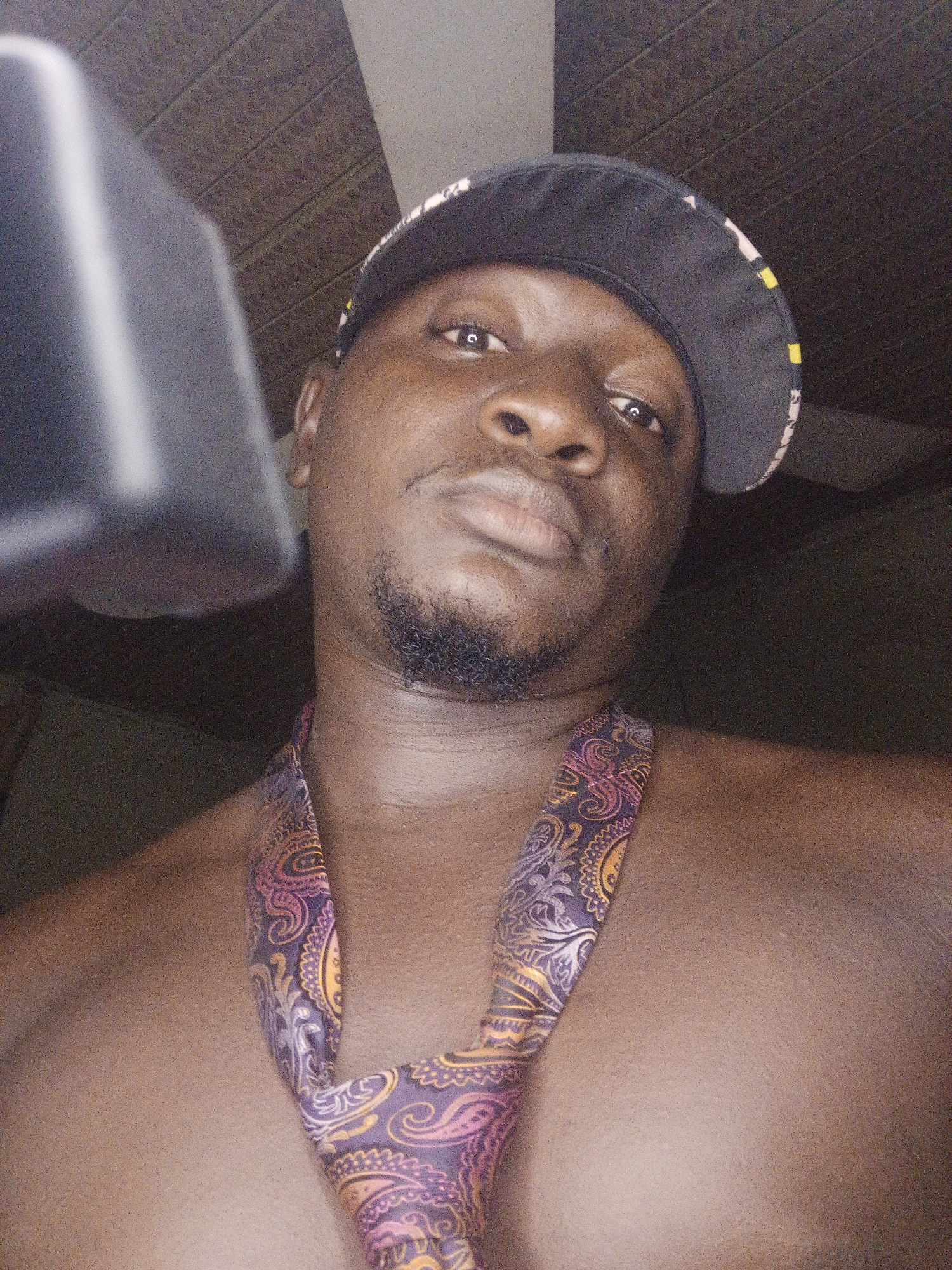 A man wearing a black baseball cap and a patterned tie around his neck, with his chest exposed, is pictured in a close-up, waist-up shot. The man has a goatee and is looking towards the camera with a neutral expression. The background consists of a textured ceiling and a dark wall, suggesting an indoor setting. The image appears to be taken in Akwanga, Nigeria, given the information provided.