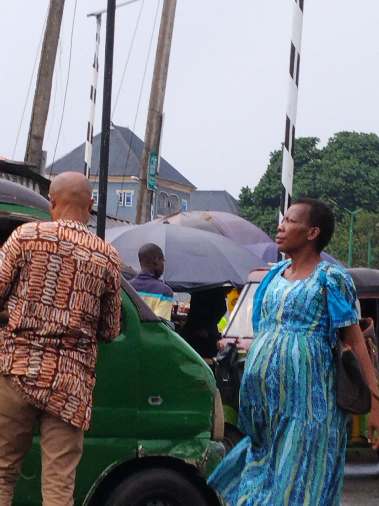 The image shows a street scene in Aba, Nigeria. In the foreground, a bald man wearing a patterned shirt and tan pants stands next to a green vehicle. A woman in a blue patterned dress and carrying a shoulder bag is visible to the right of the vehicle, looking upwards and to the left. Umbrellas are open in the background, suggesting it is raining. Other people are seen in the background, partially obscured by the umbrellas and vehicles. Overhead power lines and black and white striped posts are also visible. A building with a dark roof is in the midground behind the people. The general impression is a typical urban street scene in Aba, Nigeria, with pedestrian and vehicle traffic.
