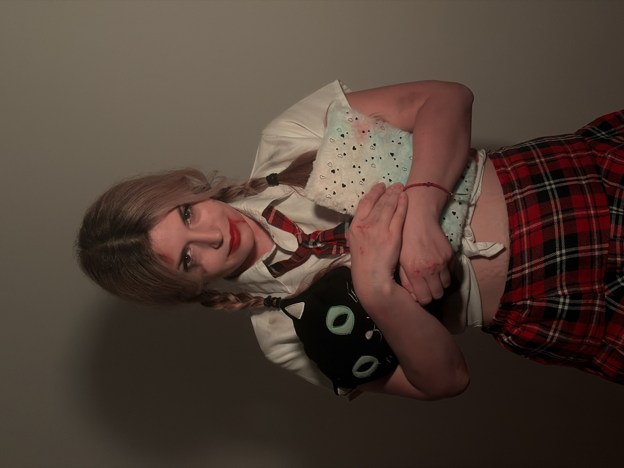 This image, captured in Budva, Montenegro, features a young woman posing indoors against a plain, dark gray wall. The photograph is taken from a tilted perspective, possibly indicating a reclined position or a stylistic choice. The woman looks directly at the camera with an intense or serious expression.
She has light brown hair styled in two braids, and her face is adorned with bold red lipstick and several red smudges or marks, particularly above her left eyebrow and around her mouth, which could suggest a dramatic makeup application or simulated injury. Similar red marks are visible on her hands. She is dressed in a schoolgirl-inspired outfit: a white collared shirt, tied at the waist to reveal her midriff, and a red and black plaid skirt. A matching plaid tie is loosely worn around her neck.
She holds two plush toys against her body. One is light blue with a pattern of black hearts, and the other is a black cat-shaped toy with large green/teal eyes and a small white triangle on its ear. The setting appears to be an indoor space with dim, artificial lighting, creating shadows that add to a potentially dramatic or slightly unsettling mood. There is no visible text in the image.