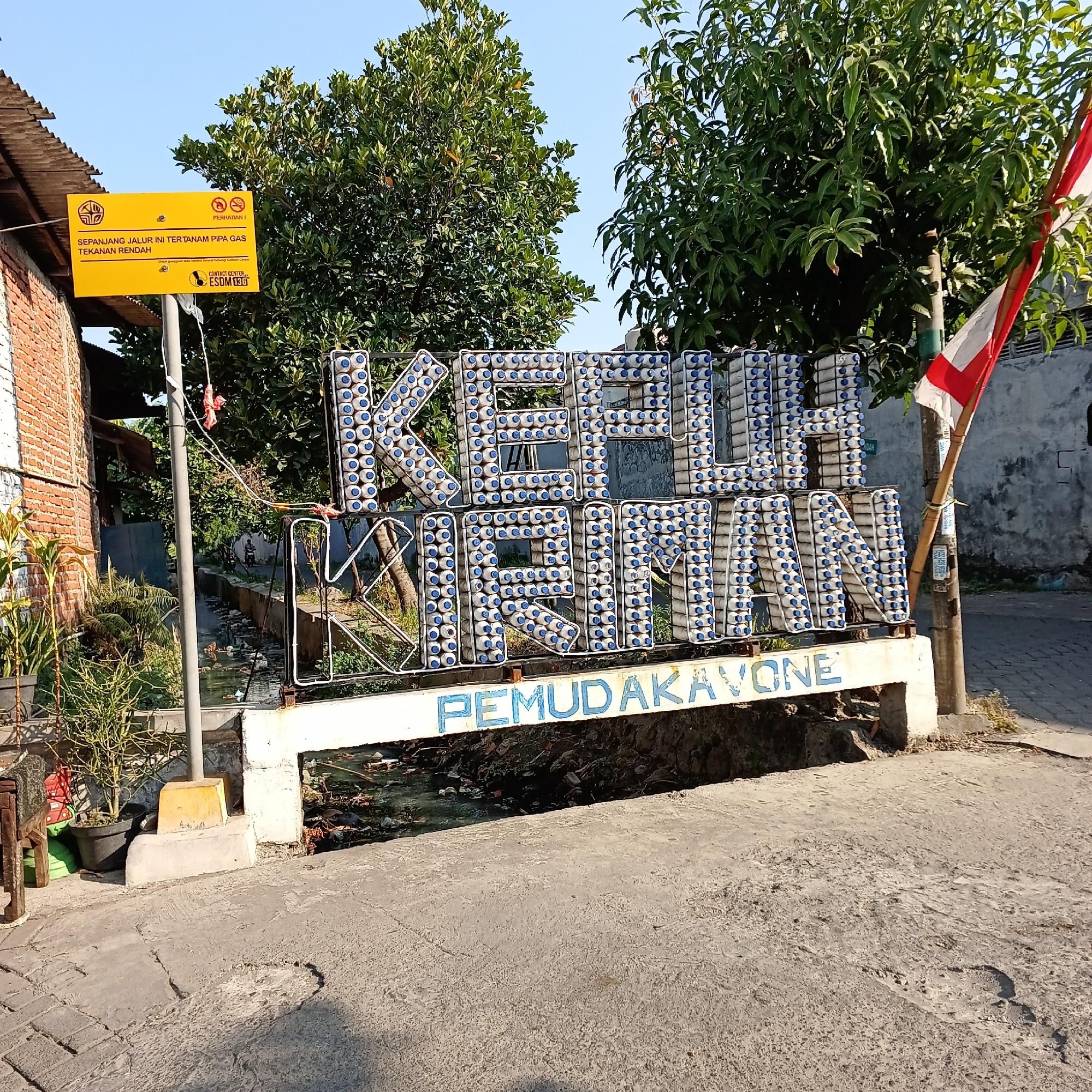 The image depicts a street scene in Wedoro, Indonesia. Dominating the foreground is a large, three-dimensional sign made of white plastic bottles with blue caps, spelling out "KEPLIHAIAN". Below this, a white banner with blue lettering reads "PEMUDAKAVONE".
To the left of the sign, a yellow warning sign is mounted on a pole. This sign has text in Indonesian, indicating "SEPONJANG JALUR INI TER TANAM PIPA GAS TEKANAN RENDAH" (This entire path is a low-pressure gas pipeline). It also includes a symbol for "PERHATIAN" (Attention) and icons depicting a smoking prohibition and an open flame prohibition, along with contact information for "ESDM139". A brick building and some potted plants are visible behind this sign.
A small canal or ditch runs behind the "PEMUDAKAVONE" banner, containing murky water and debris. On the right side of the image, a wooden pole holds an Indonesian flag, half red and half white, and some smaller signs. Trees with green foliage are present in the background, and the sky is clear and blue. The ground in the foreground is a rough, textured concrete surface with some cracks and damage. A person on a motorcycle is visible in the middle distance, on the road behind the canal.