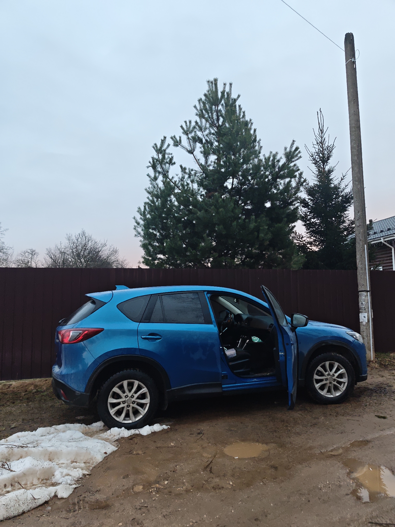 The image shows a blue Mazda CX-5 parked on a muddy, slushy ground in the city of Свистуха (кузяевский С/о), Russia. The car's driver-side door is open. Behind the car is a dark brown fence. A tall pine tree and a telephone pole stand near the fence. Patches of melting snow are visible on the ground. The sky is overcast, suggesting a cold and damp day.