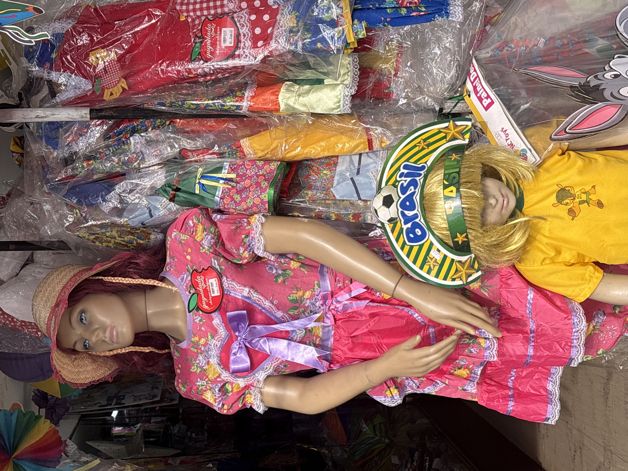 The image shows a street stall in São Paulo, Brazil, displaying various traditional Brazilian Festa Junina costumes and decorations. In the foreground, a female mannequin is dressed in a vibrant pink floral dress with a straw hat, complete with braided hair. Beside her, a smaller mannequin wears a yellow t-shirt with a Brazilian-themed headband featuring a soccer ball and the word "Brasil." The background is filled with racks of clothing, some of which are encased in plastic, showcasing a variety of colorful patterns and styles. The stall appears to be open to a street, with glimpses of other stalls and general street activity in the blurred background. The overall impression is one of a bustling marketplace, likely specializing in celebratory attire for local festivals.