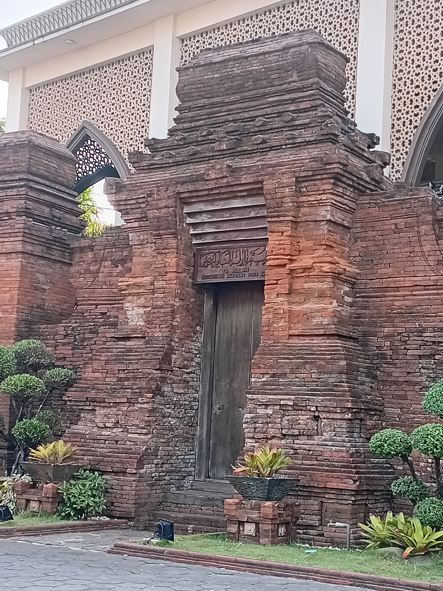 The image shows an ancient brick gate with intricate carvings above a wooden door. The gate is constructed from weathered red bricks, with layers of stepped stonework forming a tiered structure. To the left and right of the gate are ornamental trees and potted plants. The background reveals the facade of a white building with a patterned lattice screen and arched windows. The scene is set in a courtyard or garden area, with paved pathways and manicured grass. The location is identified as the Menara Kudus Mosque, in the village of Loram Kulon, Kudus Regency, Central Java, Indonesia. The time of day appears to be daytime, with natural light illuminating the structure. There are no people visible in the image.