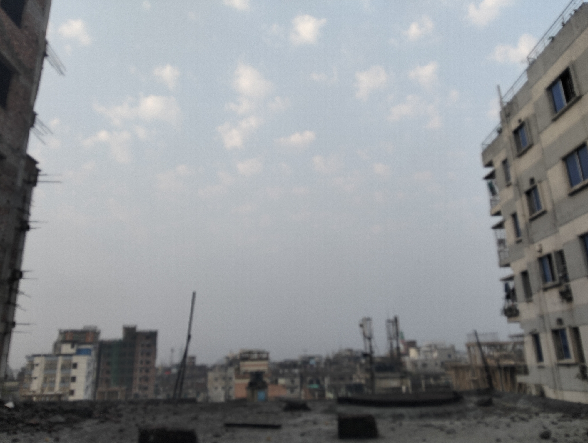 The image, taken from a rooftop in Dhaka, Bangladesh, shows a cityscape under a hazy, cloudy sky. In the foreground, the rough, unfinished surface of the rooftop is visible. To the left, there is a building under construction. To the right, a completed apartment building with windows and balconies. The background contains a mix of low-rise and mid-rise buildings, antennas, and other urban infrastructure. The scene is out of focus, but the skyline appears dense and typical of a large urban area. No people are clearly discernible in the image.
