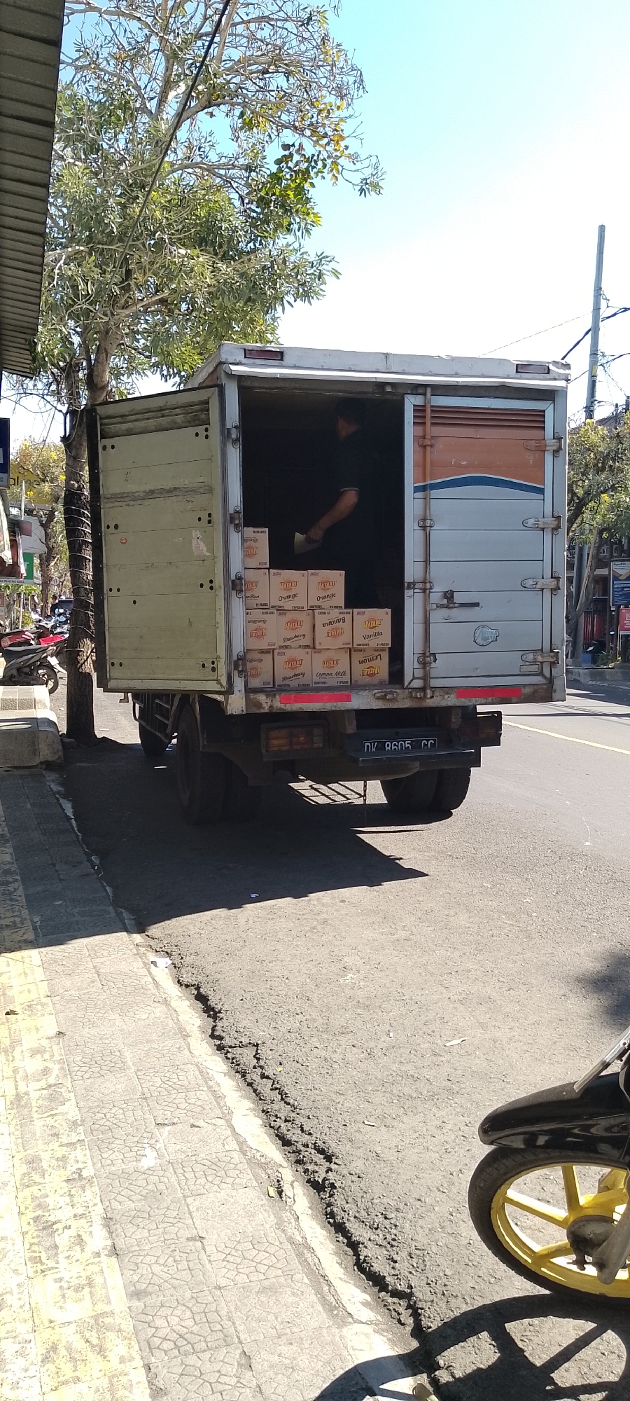 An open-backed truck is parked on the side of a road in Pekutatan, Indonesia, with its rear cargo area filled with boxes of beverages. A person, dressed in a dark shirt and pants, is inside the truck, partially obscured by the opening, appearing to be in the process of organizing or unloading the cargo. The boxes, predominantly cardboard, are stacked neatly and feature labels indicating various fruit flavors such as "Orange," "Banana," "Vanilla," "Lemon," and "Strawberry." The truck itself is a light gray color with some orange and blue detailing on the rear doors. A license plate visible on the back of the truck reads "DK 8605 CC". The truck is positioned next to a sidewalk with a patterned paving and adjacent to a tree with sparse foliage. In the background, other vehicles, including a motorcycle, are visible, suggesting a moderately busy street scene. The overall environment is outdoors, under a clear blue sky, with sunlight casting shadows on the road and sidewalk.
