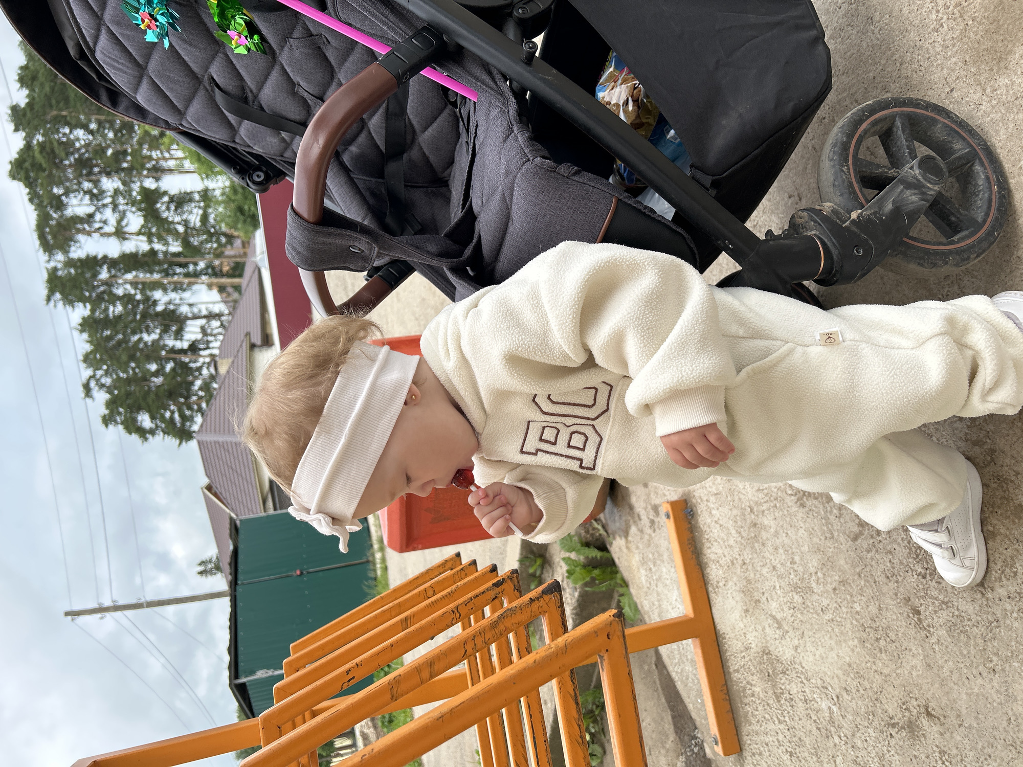 In the city of Azinsky, Russia, on a cloudy day, a young child, likely a toddler, stands outdoors next to a dark gray stroller. The child is wearing a cream-colored fleece jumpsuit with the letters "BC" embroidered on the chest and a white headband. They are holding a red lollipop to their mouth. The stroller has a brown handlebar and two bright pink straws protruding from its side. Behind the child and stroller, there is an orange metal structure with horizontal bars, possibly part of a playground or barrier. Further in the background, a green metal shed, a red container, and residential buildings with tiled roofs are visible, all set against a backdrop of tall pine trees and an overcast sky with power lines. The ground is paved with concrete.