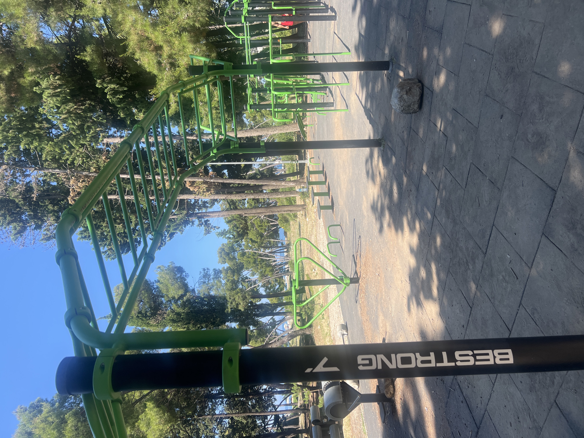 This is a medium-shot, outdoor, eye-level, full-color photograph taken on a bright, sunny day in the city of Bar, Montenegro. The image depicts an outdoor fitness park, likely located near the coast, as evidenced by the glimpse of the Adriatic Sea and a beach in the background. The fitness equipment, primarily green and black, is arranged on a dark gray paved surface. Prominently featured is a monkey bar structure with numerous horizontal bars suspended above the ground, supported by vertical poles. To the left, a black pole with white lettering reads "BESTRONG" vertically. Various other exercise apparatus, including what appear to be dip bars and benches, are visible throughout the park. The surroundings are characterized by numerous tall, slender trees with lush green foliage, casting dappled shadows on the ground. The sky is a clear, vibrant blue. A single, small, grey rock rests on the paved ground in the foreground. The presence of a person in a red shirt, partially visible on the far right, suggests the park is in use. The overall scene conveys a sense of a well-maintained public recreational area.