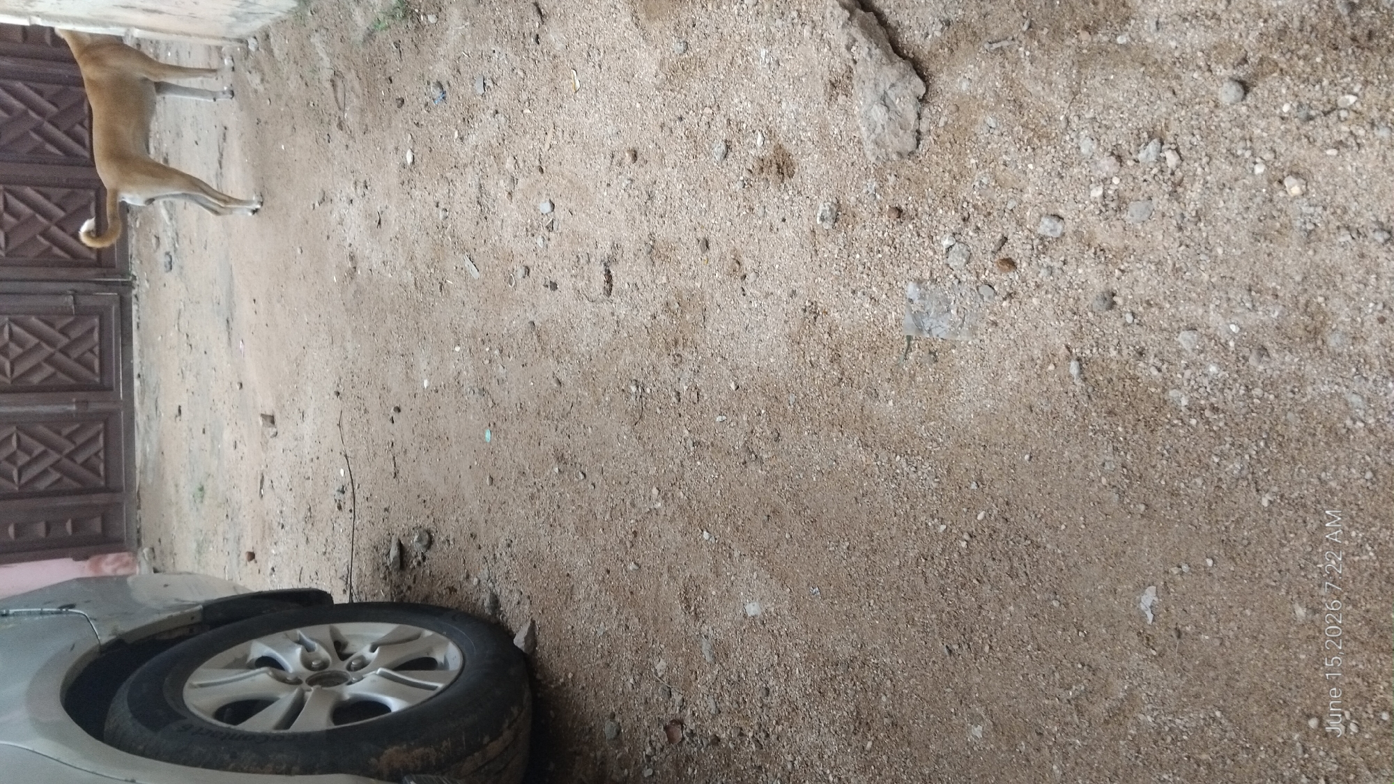 The image captures a ground-level view of an outdoor scene, likely a pathway or yard, composed of dirt and gravel. In the left foreground, a portion of a silver car is visible, specifically its rear wheel and lower body panel. The wheel features a multi-spoke silver alloy rim. Further into the background, a light brown dog is standing on the dirt surface, facing away from the camera and towards a closed, brown gate. The gate appears to be made of metal with a decorative, geometric pattern. The surrounding environment is predominantly dirt, interspersed with small pebbles and scattered debris. A portion of a building's wall, a light tan color, is visible on the far right edge of the frame. The overall impression is one of an everyday outdoor setting, possibly a residential area in Howayi, Nigeria. The timestamp "June 15, 2026, 7:22 AM" is overlaid on the bottom left corner of the image.