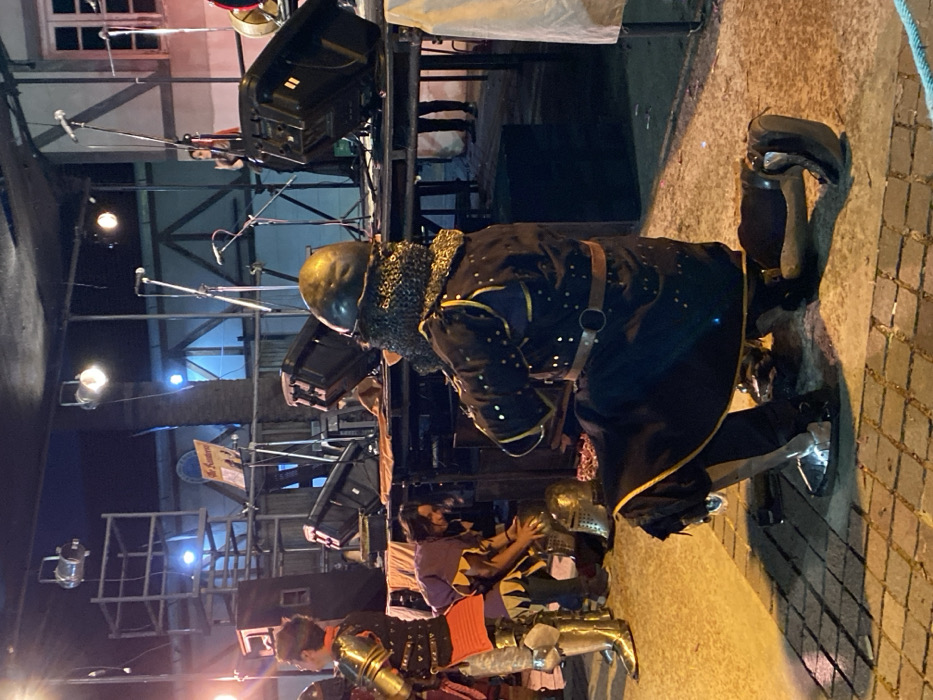 The image depicts an outdoor medieval fair or reenactment event, identified by signage as "El Konterie," within the city of Buenos Aires, Argentina, at night.
In the foreground, a male individual in medieval-style armor kneels on his right knee, viewed from the rear-right. He wears a dark tunic adorned with gold accents, a leather belt, metal gauntlets, full leg armor including sabatons, and a metal helmet with a visible chainmail coif or aventail. The ground beneath him is paved with light-colored square bricks.
To the left, another male individual stands, partially visible, wearing golden full plate armor on his legs and arms over a dark red or orange garment. Further left and slightly behind, a third male individual, dressed in a purple and yellow patterned tunic, kneels or bends over, appearing to handle one of two visible metal helmets resting on the ground.
In the midground, an elevated stage structure is visible, equipped with multiple large black stage monitors or speakers, microphones on stands, and various stage lights mounted on scaffolding. A partially visible sign with "El Konterie" is positioned on the stage. On the far right of the stage, a female individual is partially visible, standing near a microphone stand.
The background features a tall palm tree and a building exhibiting exposed timber framing and illuminated windows, indicative of a specific architectural style. The scene is illuminated by a mix of warm artificial lights in the foreground and cooler lights in the background.