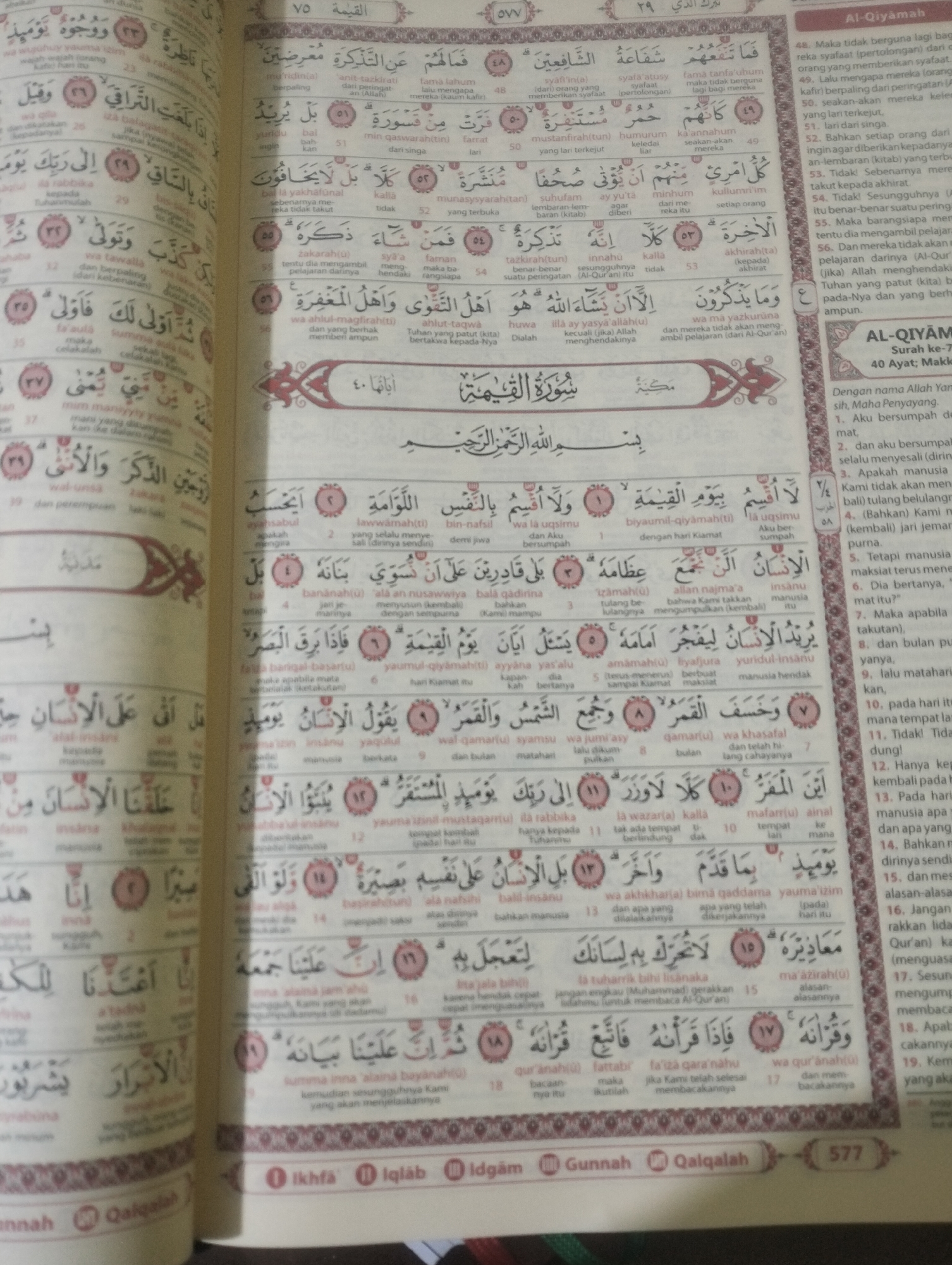 The image shows an open Quran, likely a personal copy, displaying Arabic script with accompanying translations and Tajweed rules. The book is lying flat, with its pages slightly splayed open, revealing text and some visual elements like decorative borders or chapter headings. The lighting suggests it's indoors, possibly in a home or a place of worship, given the nature of the object. No people are visible in the image, and no specific location in Dumai City, Indonesia, can be identified from the visual information provided. The focus is solely on the Quranic text and its presentation.