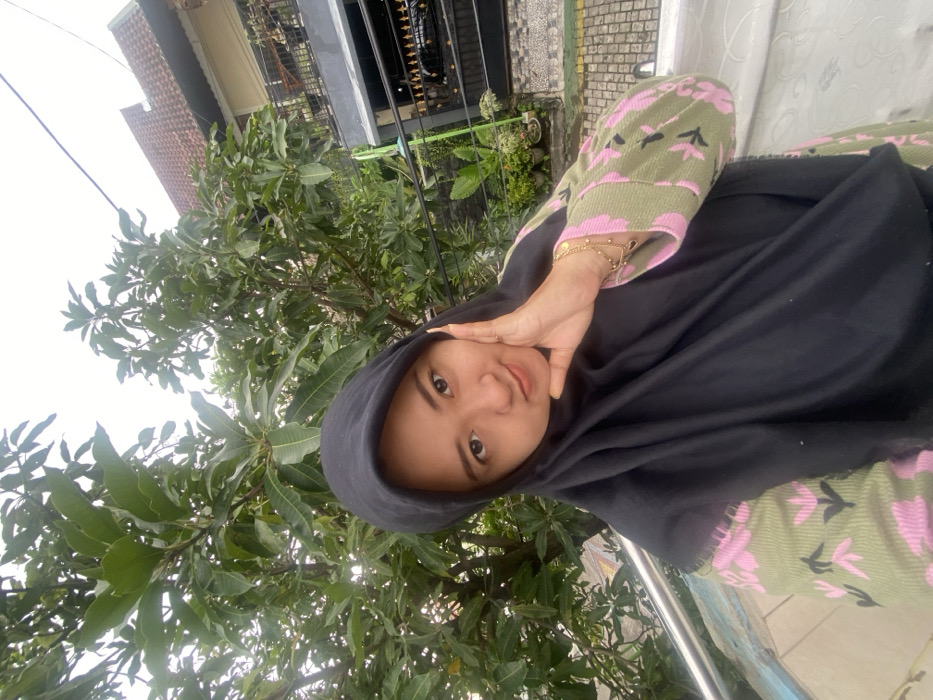 A young woman wearing a black hijab and a green and pink patterned top poses outdoors. She is close to a lush tree with large, green leaves. Her hand is raised near her face, showcasing a bracelet. The background features a building with a gray facade and a sky that appears overcast. The photograph is taken at an angle, presenting a tilted perspective.