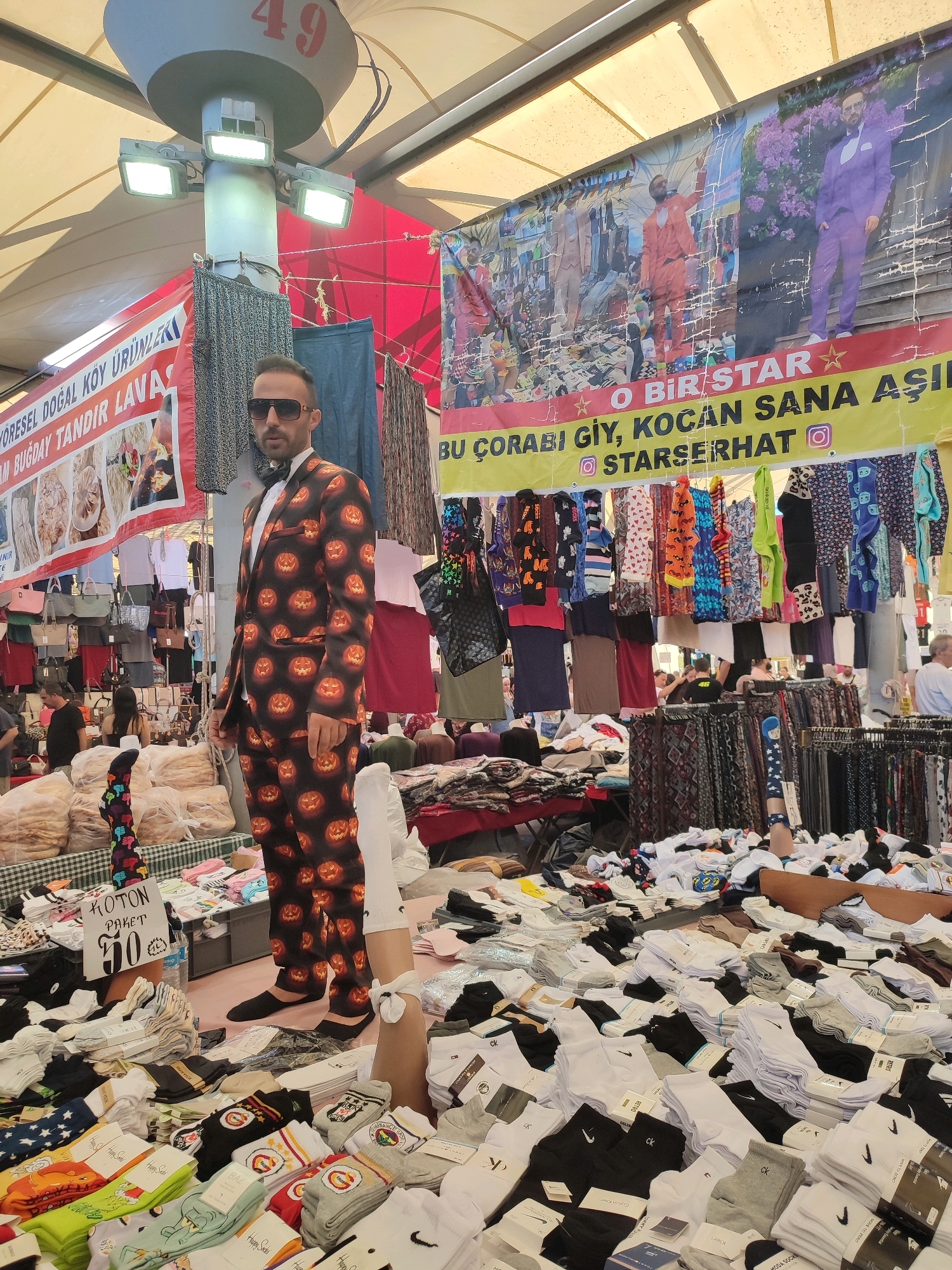 In the Kadıköy district of Istanbul, Turkey, a man stands in a market stall wearing a Halloween-themed suit patterned with jack-o'-lanterns. He is also wearing a white shirt, a black bow tie, and sunglasses. The stall displays a variety of socks, with stacks of white, black, and gray socks prominently featured. In the background, a large banner displays colorful advertisements for clothing and mentions "O BIR STAR" (You are a star) and "KOCAN SANA ASİK OLACAK" (Your husband will fall in love with you). The market appears to be outdoors, with a tented roof providing shade, and other vendors and customers are visible in the background.