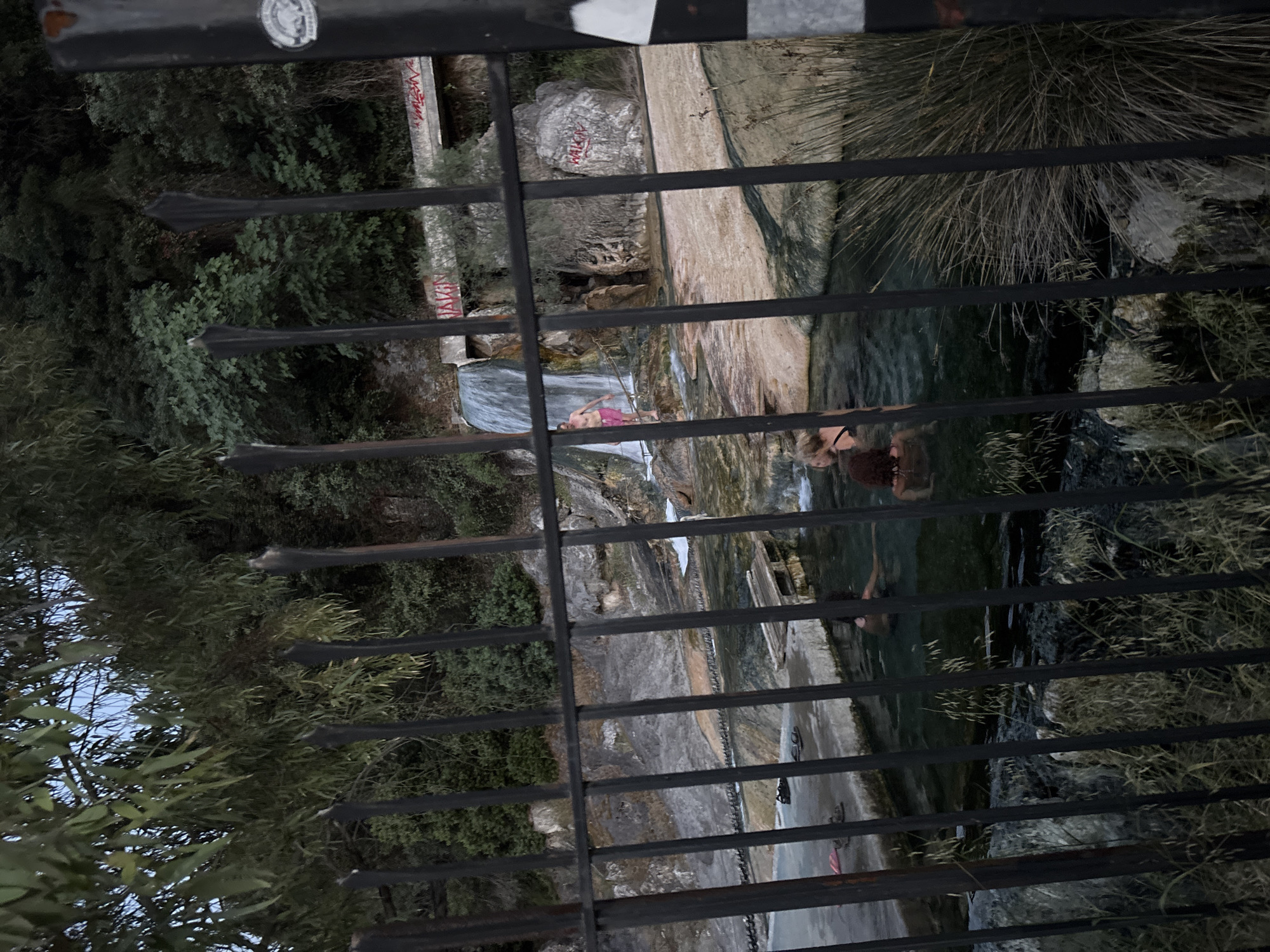 The image shows a fenced-off view of the Thermopyles Hot Springs in the Municipal Unit of Lamia, Greece. The viewpoint is through a metal fence, with greenery and trees visible on the left and behind the fence. The springs consist of multiple pools, with people swimming and relaxing in them. Water flows over a series of rocks, creating small waterfalls. Graffiti is visible on some of the rocks.