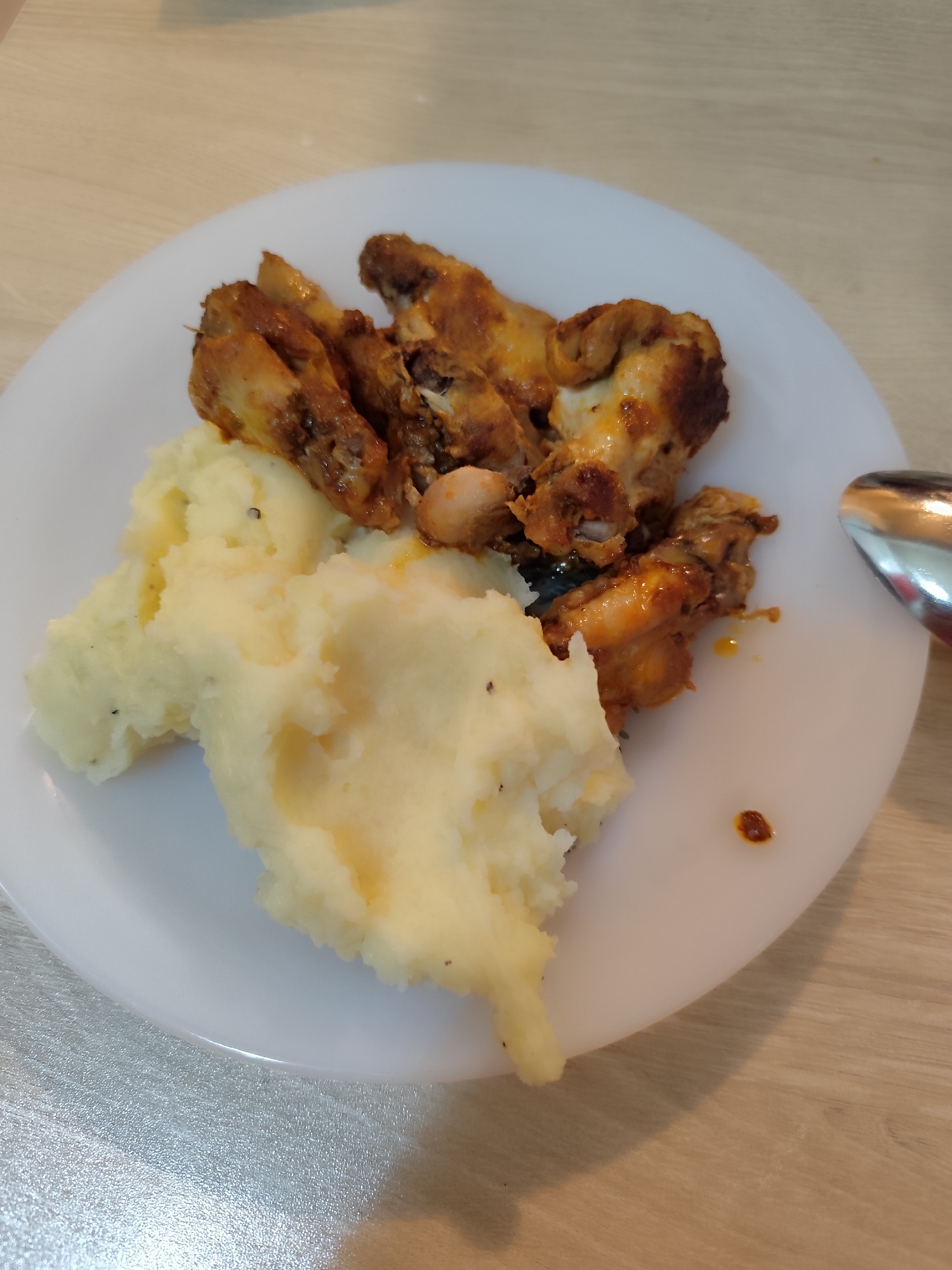 On a white plate, there is a serving of mashed potatoes alongside several pieces of cooked chicken. A spoon rests on the right edge of the plate. The plate is placed on a wooden surface. The location is not discernible from the image.