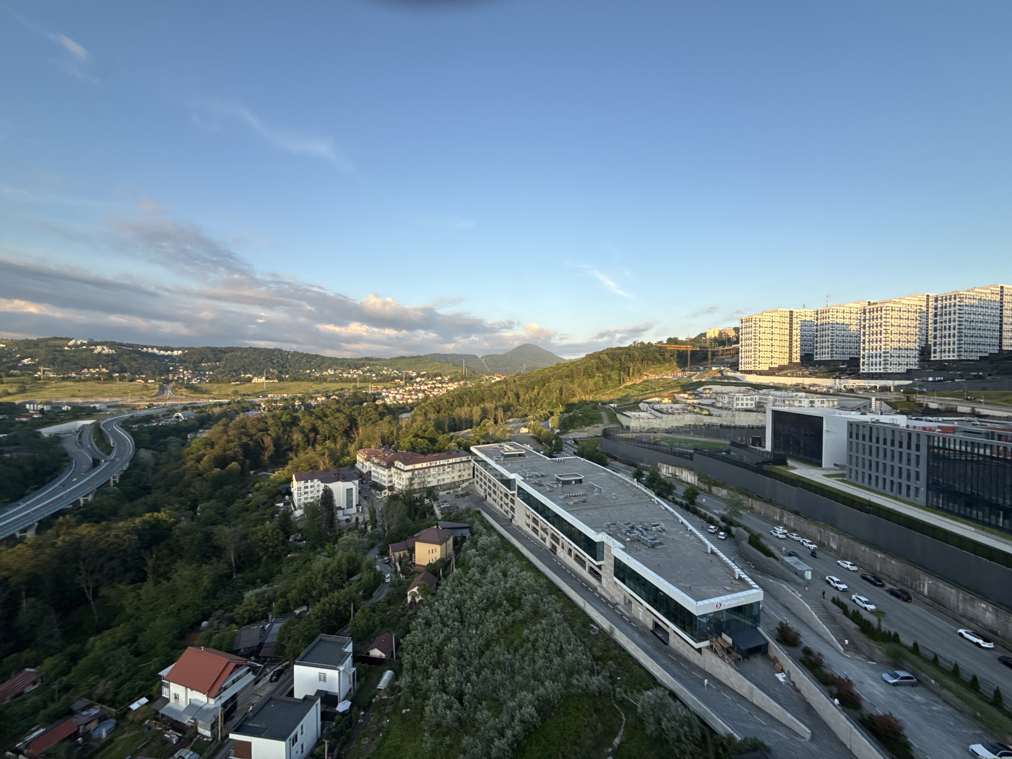 This is an aerial, wide-angle photograph taken at a low angle during the golden hour in Sochi, Russia. The scene captures a blend of urban development and natural landscape under a clear blue sky with scattered clouds.
In the foreground, a modern, long, multi-story building with a white facade and green-tinted windows stretches across the frame. This building appears to be part of a commercial or residential complex, with its roof featuring ventilation units and other equipment. A road runs alongside this building, with several cars parked and driving on it.
Further to the right, a dense cluster of identical, multi-story apartment buildings, with white facades and dark windows, are visible. These buildings are situated on a hillside, suggesting a planned residential development. In the background, more hills covered in lush green trees extend towards the horizon, with a few scattered buildings visible among them.
To the left of the frame, a complex highway interchange with multiple levels and curving ramps is a prominent feature, indicating significant infrastructure in the area. Smaller residential houses with red and white roofs are nestled among trees on the slopes below the highway.
The overall impression is of a rapidly developing area where modern architecture is integrated into a hilly, green environment, likely on the outskirts of Sochi, given the scale of the buildings and the presence of the highway. The lighting suggests it is either early morning or late afternoon.