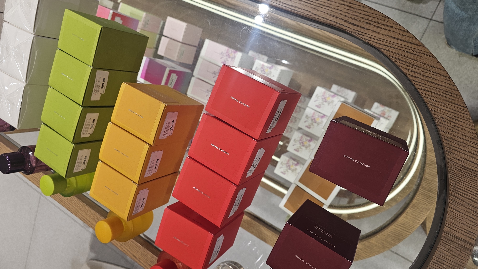 The image shows a display of stacked boxes in varying colors, likely containing perfume or scented products. The boxes are arranged in columns of green, yellow, red, and burgundy. Each box has a price tag of "399.00" visible on its side. The red and burgundy boxes are labeled "Weekend Collection" and "Cherry Temptation". The products are displayed on a reflective surface within a rounded, glass-topped table or display case with a wooden frame. The surrounding environment suggests a retail setting, possibly a shop selling perfumes or personal care products. The image does not contain explicit information indicating it is in Puebla, Mexico.