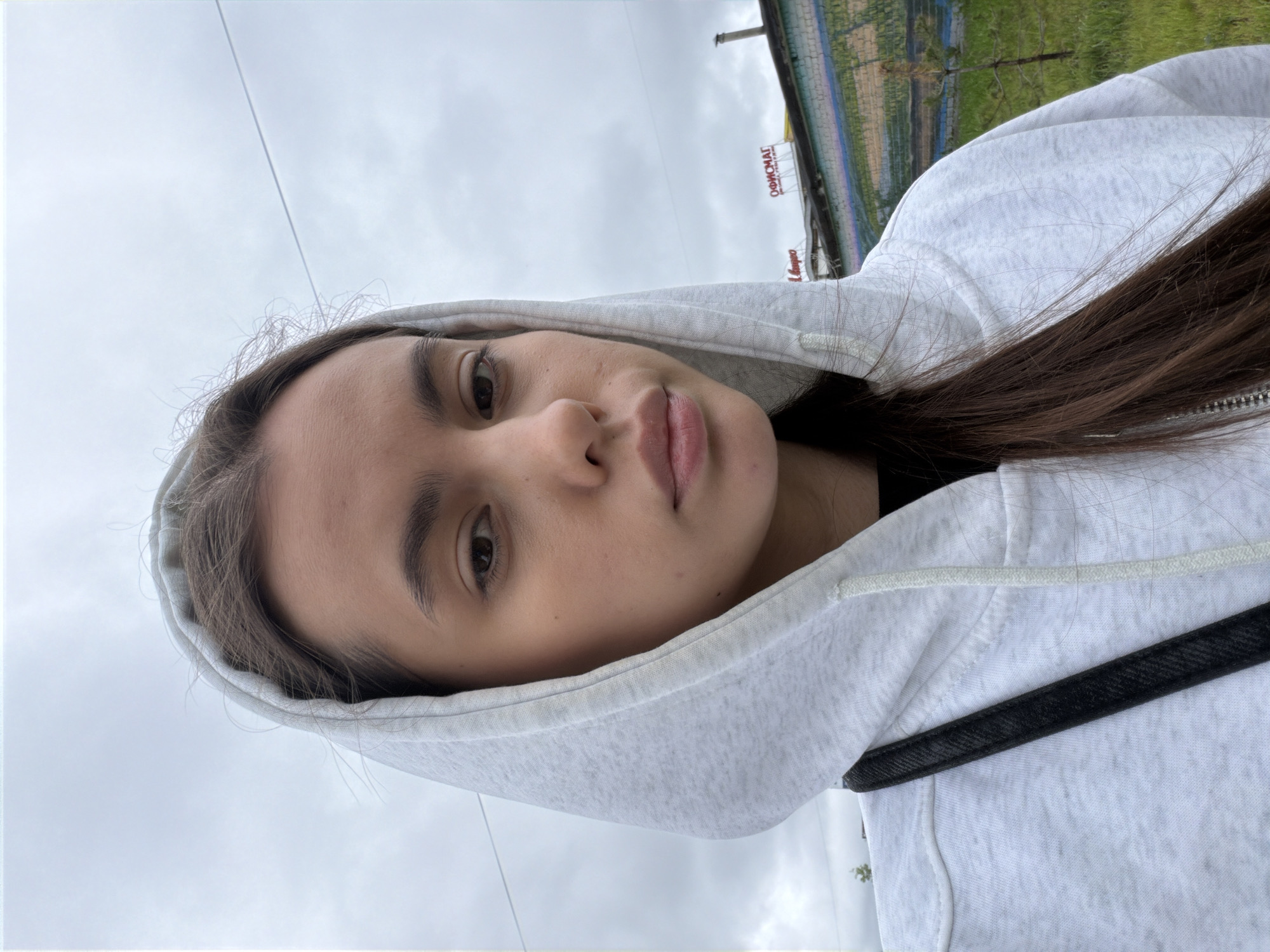 A young woman wearing a light-colored hoodie with the hood up is posing for a selfie. Her dark hair is visible from under the hood on the right side of the frame. In the background, a building with a sign written in Cyrillic reads "ОФИСМАГ", suggesting the location is likely in Russia. The image shows a cloudy sky and some greenery in the background, hinting at an outdoor environment. Based on the Cyrillic sign, the location is likely within the city of Chaikovsky, Russia, as indicated.