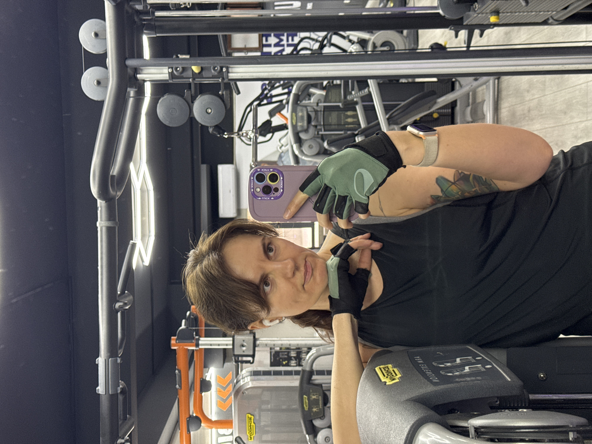 The image is a selfie taken by a woman in a gym. She is wearing workout gloves and a smartwatch, and she is holding a smartphone with a lavender case. The background shows various gym machines. There is no information in the image to confirm if the location is in the city of Budva, Montenegro.