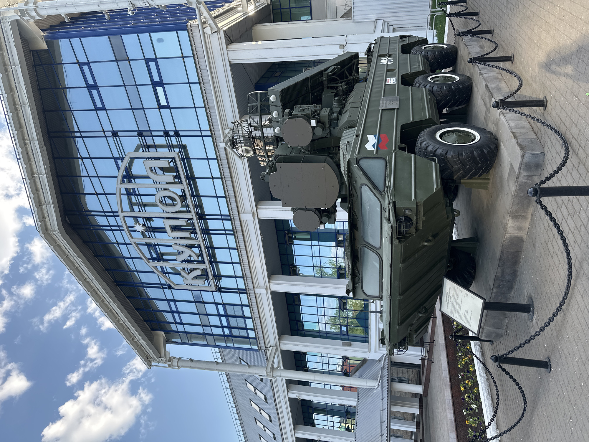 A military vehicle, specifically a Russian-made anti-aircraft missile system, is displayed outdoors in front of a modern building. The building features a reflective glass facade with white architectural columns and a large sign in Cyrillic script that reads "ROSTEH". The vehicle is olive green and has multiple large wheels. A sign with technical information is next to the vehicle. A black chain is used to rope off the vehicle from the public. The location is Izhevsk, Russia, due to the presence of the Rosteh sign, which is likely the "Kalashnikov" concern building. The sky is partly cloudy. The ground is paved.