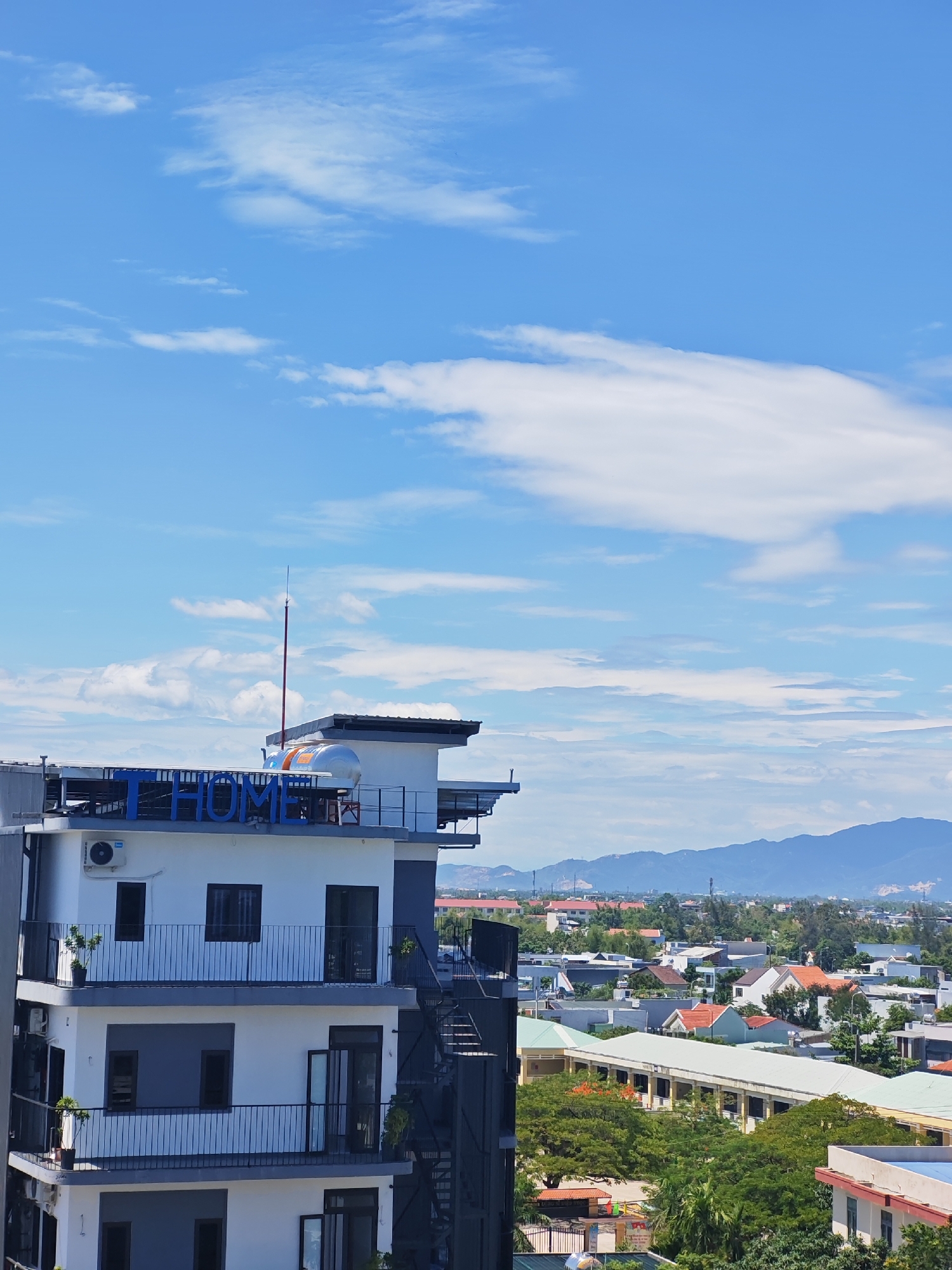 A medium shot shows a multi-story building with a sign that reads "THOME" in blue capital letters against a bright blue sky with scattered white clouds. The building has a modern design with white walls, gray accents, and black railings on balconies. There are several windows and doors visible, as well as an air conditioning unit on one of the balconies. A metal fire escape is attached to the side of the building. The building is situated in a city with other residential and commercial structures, trees, and distant mountains. The scene appears to be in Đà Nẵng, Vietnam, based on the architectural style and the presence of lush vegetation and mountainous terrain common to the region. The sky is clear, suggesting daytime with good weather conditions.