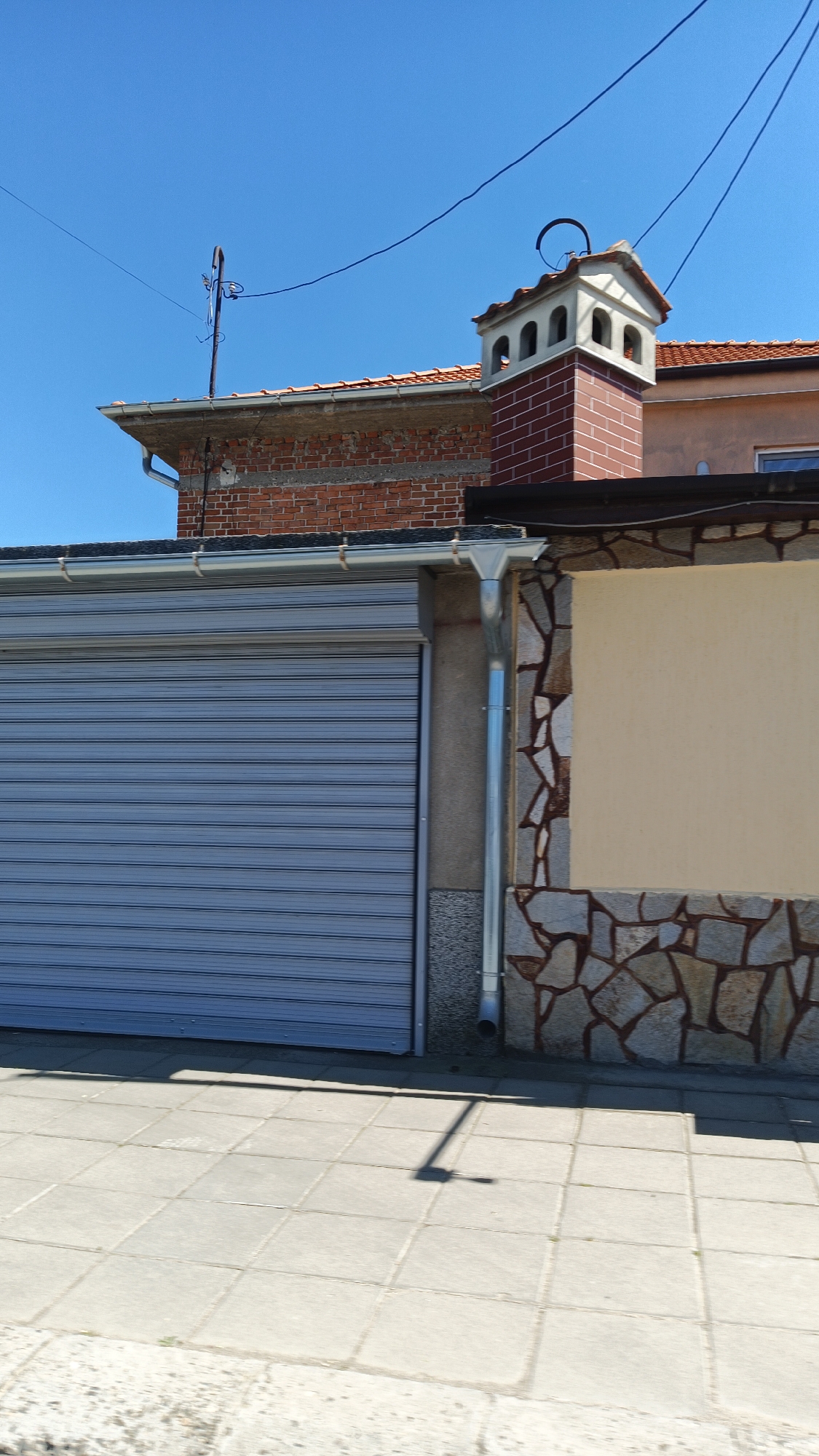 The image depicts a street scene in Milevo, Bulgaria. In the foreground is a gray tiled sidewalk. A building with a corrugated metal garage door and a decorative stone facade is visible. A brick building with a red tile roof is partially visible behind the first building. A brick chimney with a decorative top and a metal antenna are on the roof. Power lines run across the clear blue sky. There are no people visible.