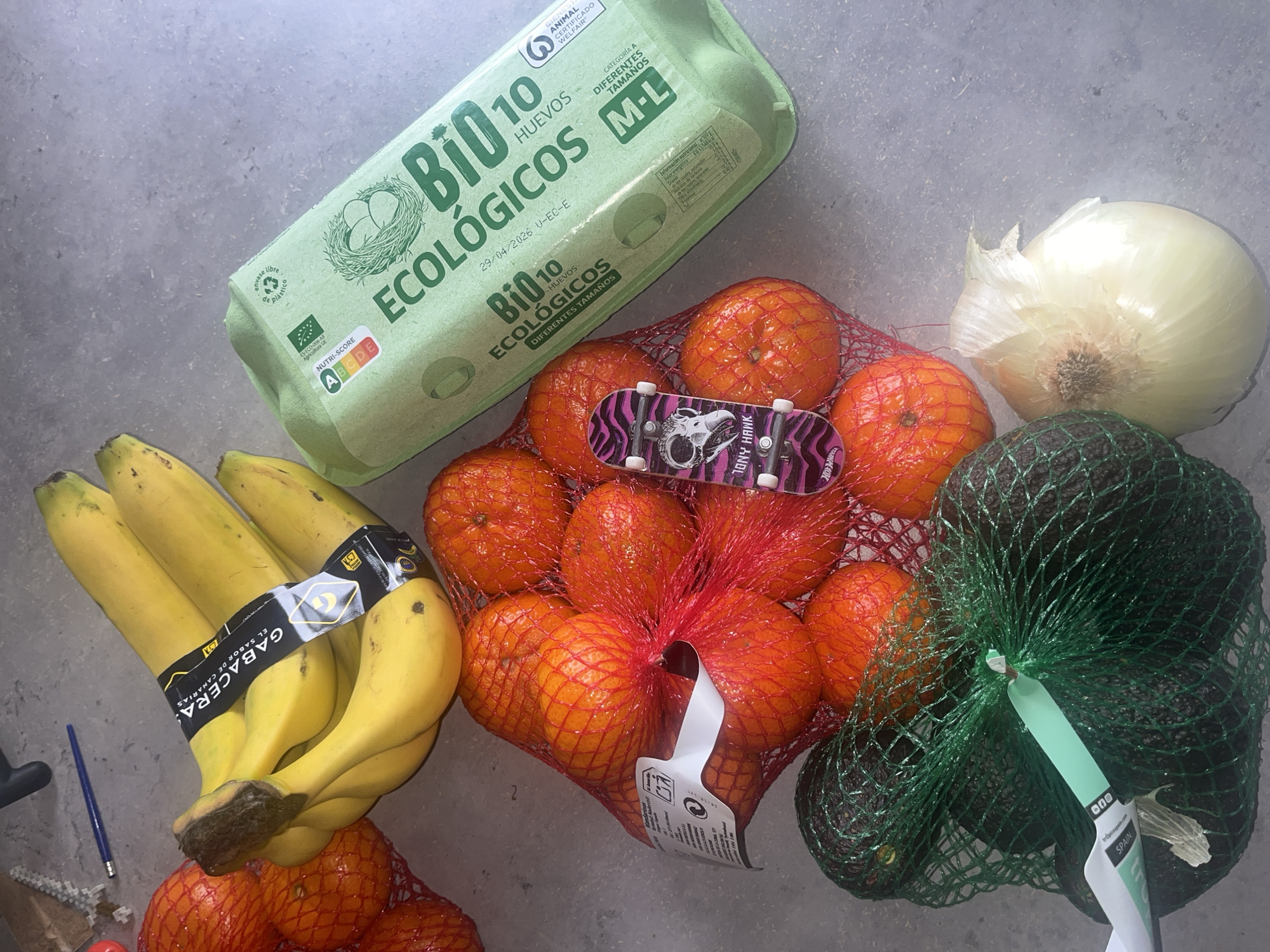 The image shows a flat lay of food items and a toy skateboard on a grey surface. The food items include a green carton of "Bio 10" organic eggs, a bunch of yellow bananas with a "Gabacera" brand label, a net bag of oranges, a white onion, and a net bag of avocados with a label indicating they are from Spain. The toy skateboard, with a purple and black design, is placed on top of the net bag of oranges. A blue pen and a small red object are located near the bananas. The image was most likely taken in l'Hospitalet de Llobregat, Spain, due to the Spanish text on the labels.