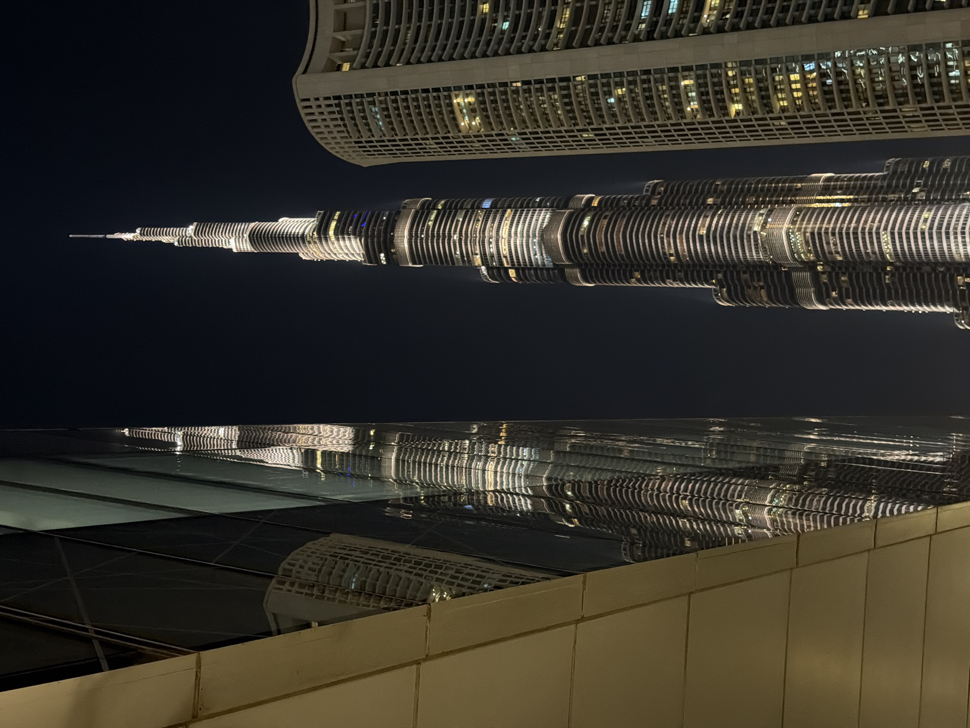 At night, a cityscape is visible in Dubai, United Arab Emirates. The Burj Khalifa, lit up, is reflected in a glass-covered surface, implying a modern architectural setting. A building with many windows appears above the Burj Khalifa and is also lit. An adjacent building’s facade is below the reflection.