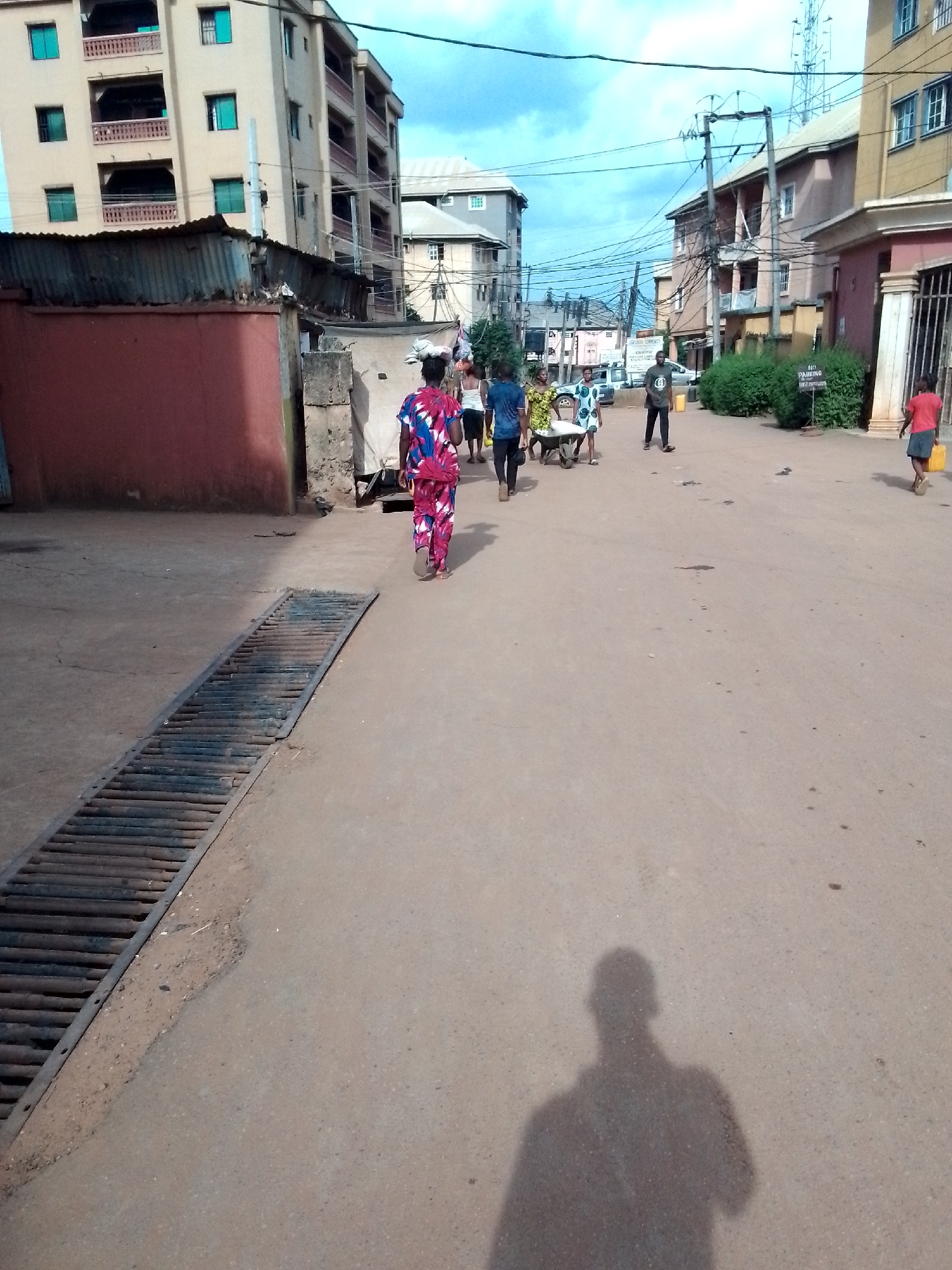 The image depicts a street scene in Onitsha, Nigeria. The street is paved, with a large metal drainage grate visible on the left side. Several people are walking along the street, some appearing to be engaged in conversation. A man is pushing a wheelbarrow. Buildings of varying heights and architectural styles line the street, including multi-story residential structures. Power lines stretch across the sky. The lighting suggests a sunny day. The shadow of the photographer is visible in the foreground.