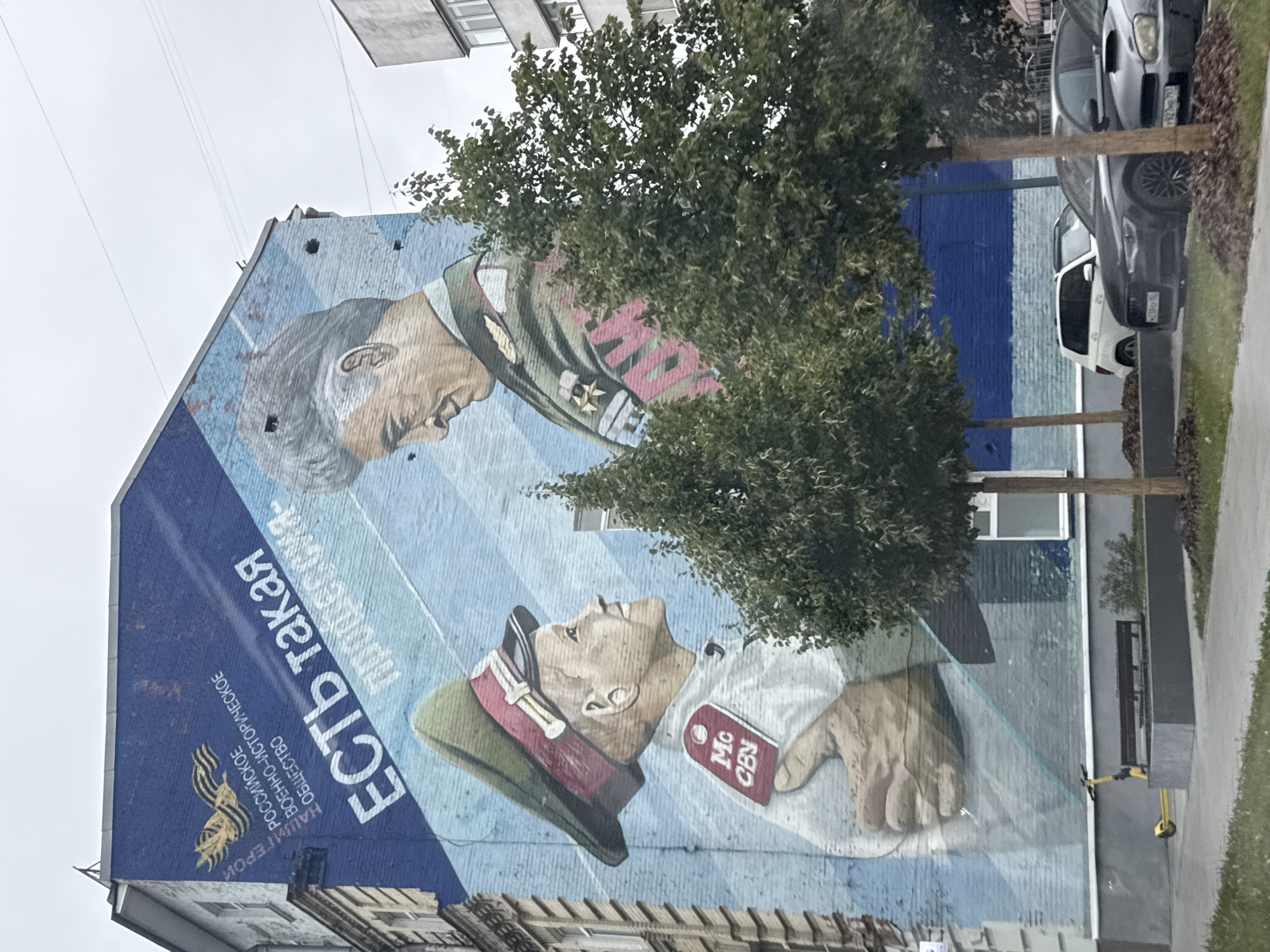 This is a photograph of a mural painted on the side of a multi-story brick building in Moscow, Russia. The mural depicts two figures in military uniforms. The larger figure on the right is an older man with gray hair, wearing a dark green military jacket adorned with medals and a red sash. The smaller figure on the left is a younger man, also in a military uniform with a green cap and red trim, looking up at the older man. The mural is predominantly blue and light blue, with some white lettering. The text in white above the younger man's head appears to read "Есть такая профессия" which translates to "There is such a profession." To the left of the figures, there is a logo with a bird and what appears to be a ribbon. In front of the building, there are several cars parked, trees, a bench, and a yellow electric scooter. The sky is overcast.