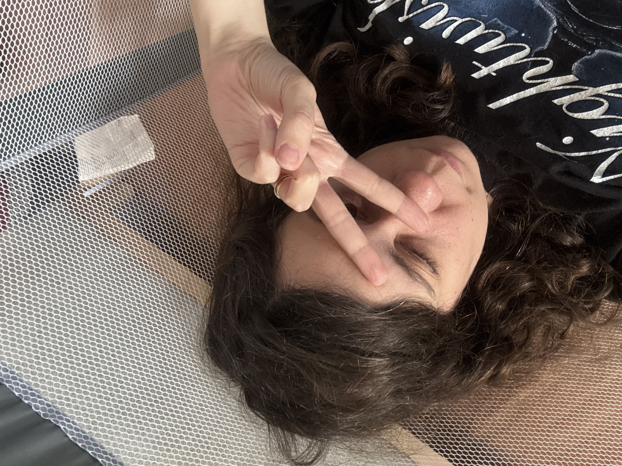 A person is lying down, with their face tilted upwards and to the right. They have dark, curly hair that falls around their shoulders. Their eyes are closed, and they are holding up their right hand to their face, forming a peace sign with two fingers extended over their forehead and cheek. A ring is visible on their ring finger. The person is wearing a dark t-shirt with white, stylized lettering that appears to read "Nightwish."
The person is positioned on a surface covered by a fine white mesh netting, which also partially obscures a wooden surface underneath. A small white tag is visible attached to the mesh in the upper right quadrant of the image. The overall environment appears to be an indoor setting, possibly a room or a portable crib, with warm lighting. The image is a close-up, taken from a high angle looking down at the person. The specific location in the city of Červená Píska, Czechia cannot be determined from the visual information provided.