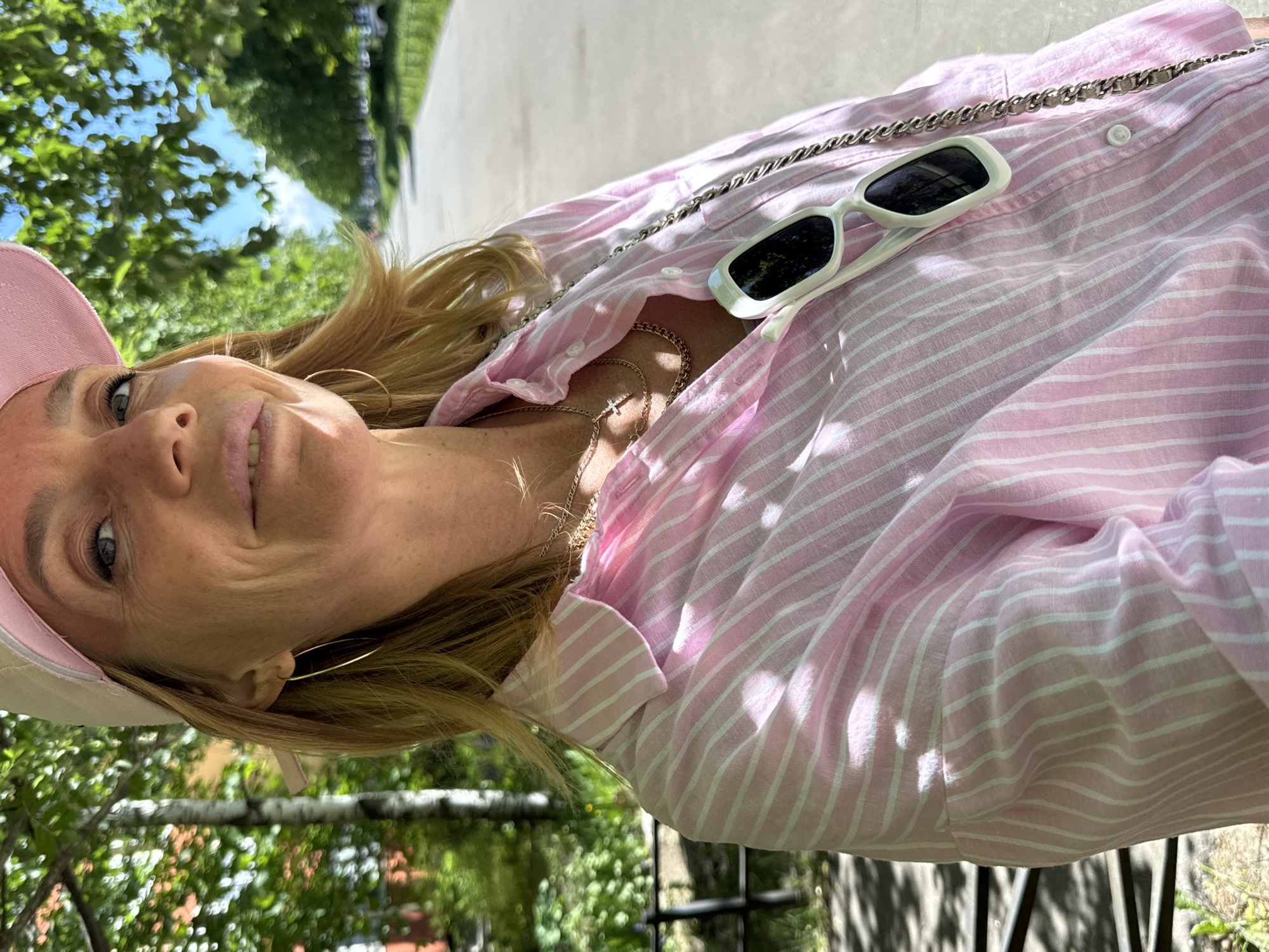 A close-up, outdoor portrait features a woman captured from a low angle, looking upwards and slightly to her left, with a subtle smile. Her blonde hair is visible, flowing around her shoulders. She wears a light pink baseball cap and a casual pink and white striped button-down shirt, partially unbuttoned to reveal gold chain necklaces, one with a small cross pendant. Large gold hoop earrings adorn her ears. White, rectangular sunglasses are perched on her shirt collar, alongside a silver-colored chain, possibly from a bag or glasses strap.
The background is a blurred outdoor scene, with vibrant green foliage and patches of bright blue sky, suggesting a sunny day in Saint Petersburg, Russia. Strong sunlight creates highlights and shadows on her clothing and hair, indicating clear weather. The overall impression is one of relaxation and enjoyment in an outdoor setting. There is no visible text in the image.