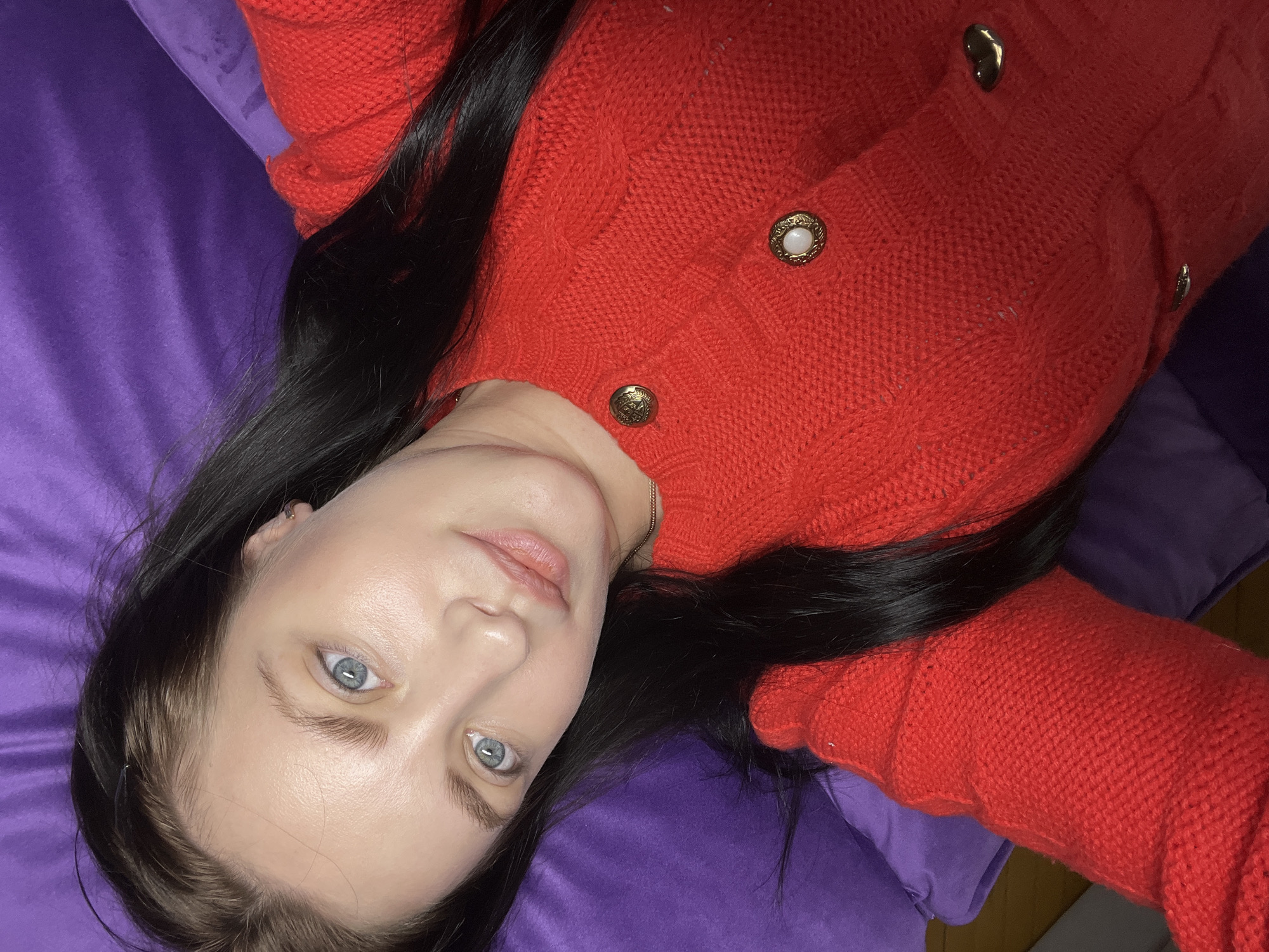 A woman with fair skin, light blue eyes, and long, straight, dark hair is lying on a purple surface, possibly a cushion or pillow. She is wearing a red knitted cardigan with gold buttons. The lighting is soft, and the background appears to be indoors. The image does not provide enough information to determine a specific location in the city of Марковский, Russia.