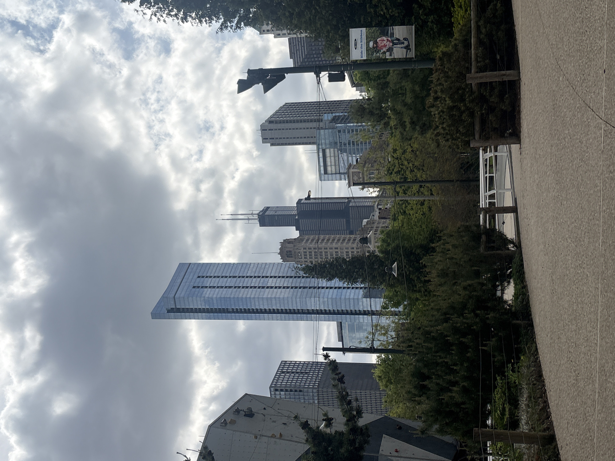 The image depicts a view of the Chicago skyline on a cloudy day. Prominent buildings include the St. Regis Chicago, the former John Hancock Center, and Willis Tower. The view is taken from ground level, looking up through trees, and partially obscuring some buildings. There is a street sign and a climbing wall in the foreground. The location appears to be in a park-like setting, potentially along the lakefront area of Chicago. There are no people visible in the shot.