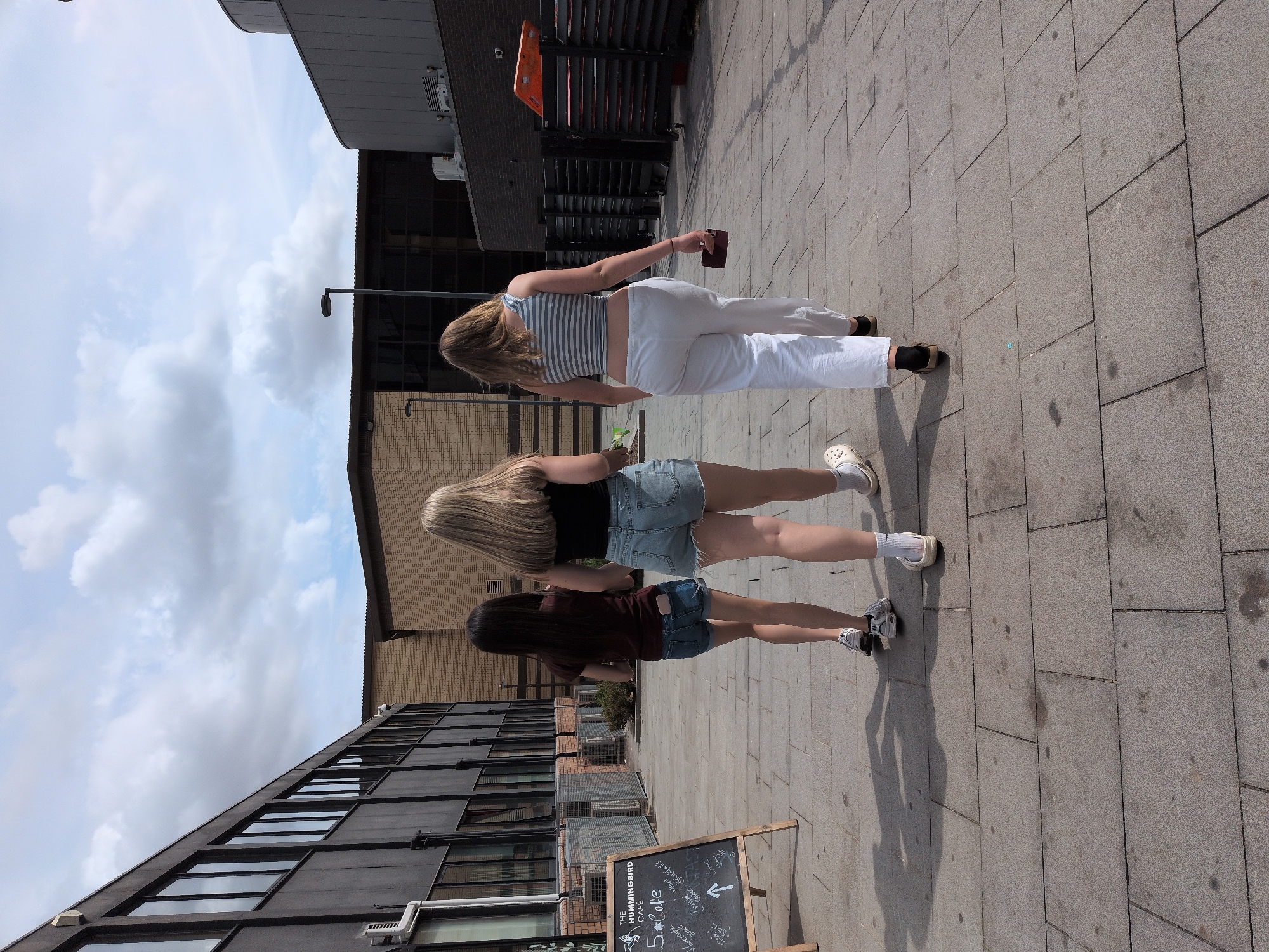 Three young females are walking away from the camera on a paved pedestrian area. The female on the left has dark hair and is wearing a dark t-shirt and denim shorts. The middle female has blonde hair and is wearing a black t-shirt and denim shorts. The female on the right has blonde hair and is wearing a striped crop top and white trousers. To the left of the image, a blackboard sign stands on an easel, advertising "The Hummingbird Cafe" with various menu items. In the background, there are buildings constructed of brick and dark metal, along with streetlights and some outdoor seating areas. The sky is cloudy. The location appears to be a modern urban environment, possibly a plaza or walkway adjacent to commercial buildings, and is identified as being in the city of Telford, United Kingdom.