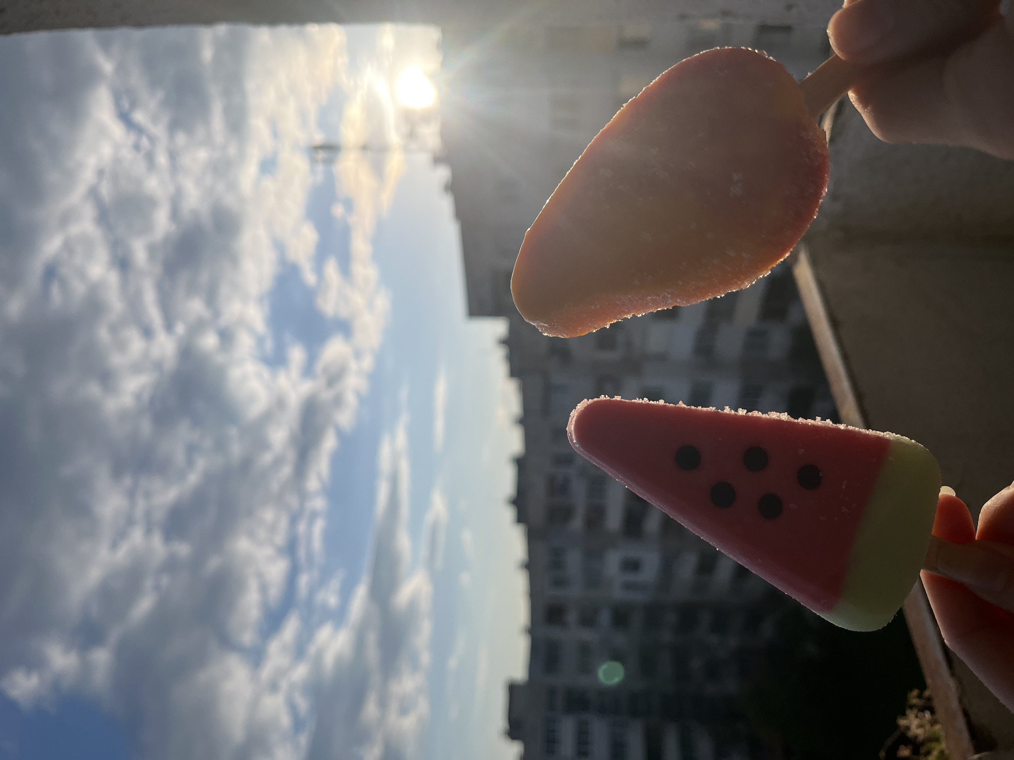 The image, rotated 90 degrees clockwise, captures two popsicles held against a bright, cloudy sky and a blurred urban background, likely Sofia, Bulgaria. The scene depicts a sunny day with distinct clouds and patches of blue sky.
In the foreground, partial views of two hands are visible, holding the sticks of two different popsicles. The upper popsicle is a solid reddish-orange, with a slightly irregular, rounded shape, and appears frosty. Below it, the second popsicle is shaped like a watermelon slice, featuring a vibrant red body with several black circular "seeds" and a light green base resembling a rind. Both popsicles have a frosty texture, suggesting they are cold and refreshing.
The sun is prominently visible in the upper part of the sky, casting bright rays and backlighting the scene, which creates a slight halo effect around the popsicles and highlights their frosty surfaces. In the distant background, silhouetted buildings of a cityscape are faintly discernible, providing context for an urban setting.
The activity suggests a person enjoying frozen treats outdoors on a warm, sunny day. No visible text is present in the image.