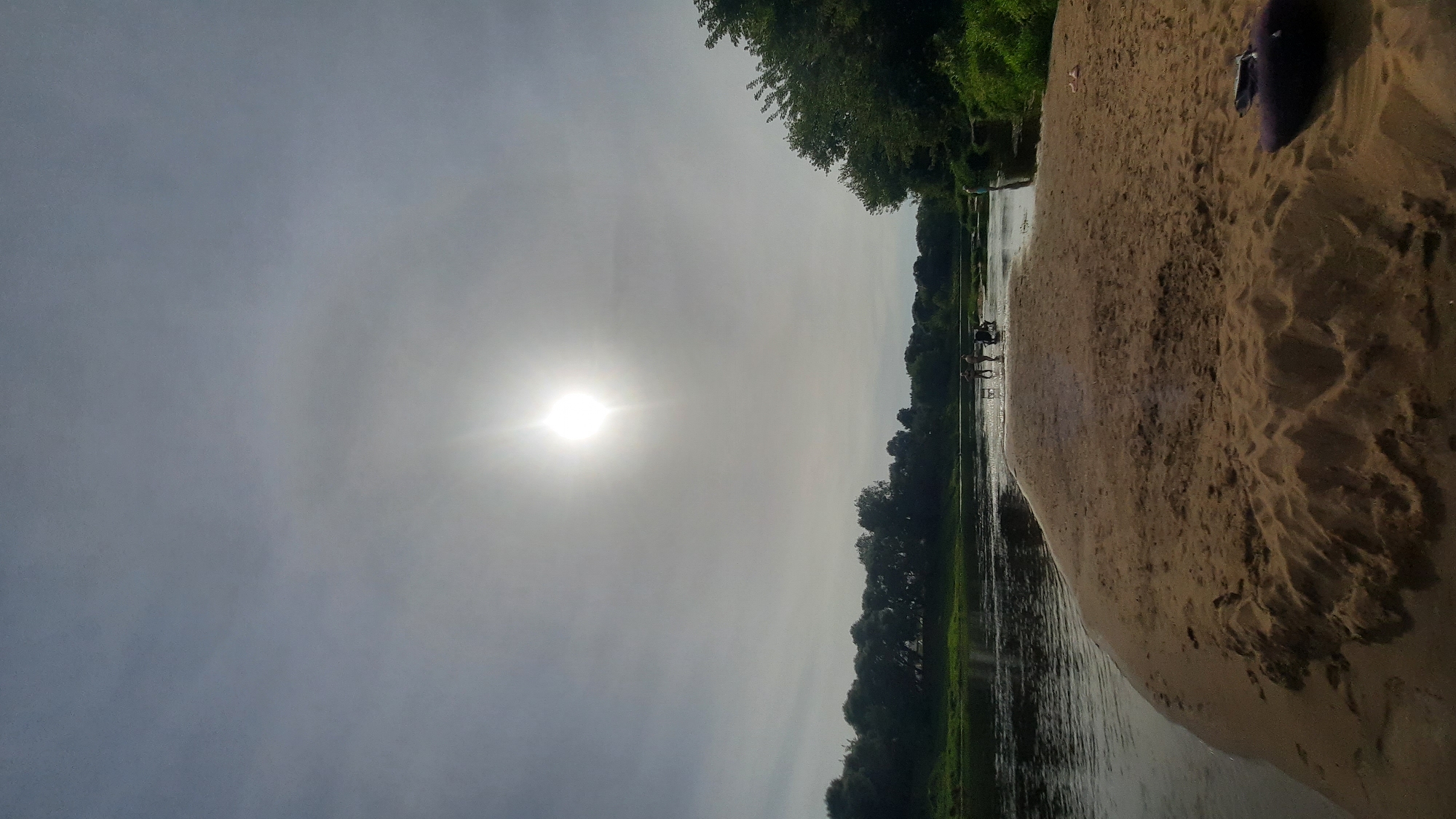 This is a vertical, eye-level, outdoor photograph taken on a sunny day at a riverbank in Shklov, Belarus. The sun is bright and high in the sky, partially obscured by thin clouds, creating a bright haze. In the foreground, a sandy beach slopes down to the water, with footprints visible in the sand. To the right, a dark object, possibly a bag or towel, rests on the sand. The river is calm and reflects the sky. On the far bank, a dense line of green trees forms the horizon, with some trees extending into the right side of the frame. In the shallow water, several people are visible. One person stands near the right edge of the frame, partially in the water. Closer to the center of the river, a group of three to four individuals are gathered, some appearing to be children. There are also what appear to be two foldable chairs or stools in the water with the group. The overall scene depicts a leisurely day at the river.