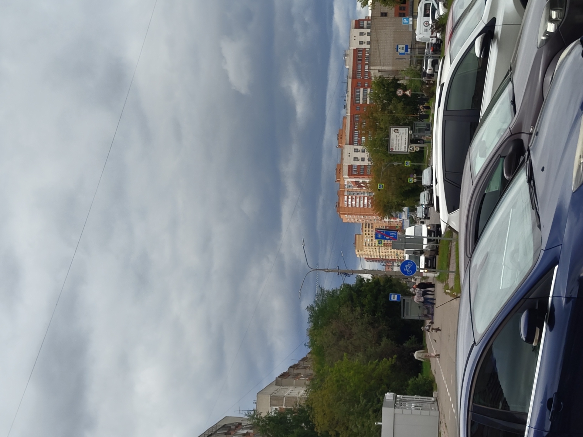 The image shows a street scene in Zhukovsky, Russia, on a cloudy day. In the foreground, the hoods and windshields of several parked cars are visible, including a dark blue car on the left, a dark gray car in the middle, and a white SUV behind it. The cars are parked on asphalt.
The middle ground features a sidewalk and a street with traffic signs and streetlights. A man in a light-colored jacket and pants walks away from the camera on the sidewalk. A child is riding a small bicycle nearby. Several other people are gathered on the sidewalk, appearing to be waiting or conversing. A blue bus stop sign is visible on a pole. A white van and other vehicles are parked on the street.
In the background, apartment buildings of varying heights and architectural styles line the street. One building is predominantly orange-red brick with white accents and many windows, while others are lighter in color. Trees with green and some autumn-colored foliage are scattered throughout the scene. Overhead, dark, heavy clouds dominate the sky, obscuring the sun. A thin power line stretches across the top of the frame.