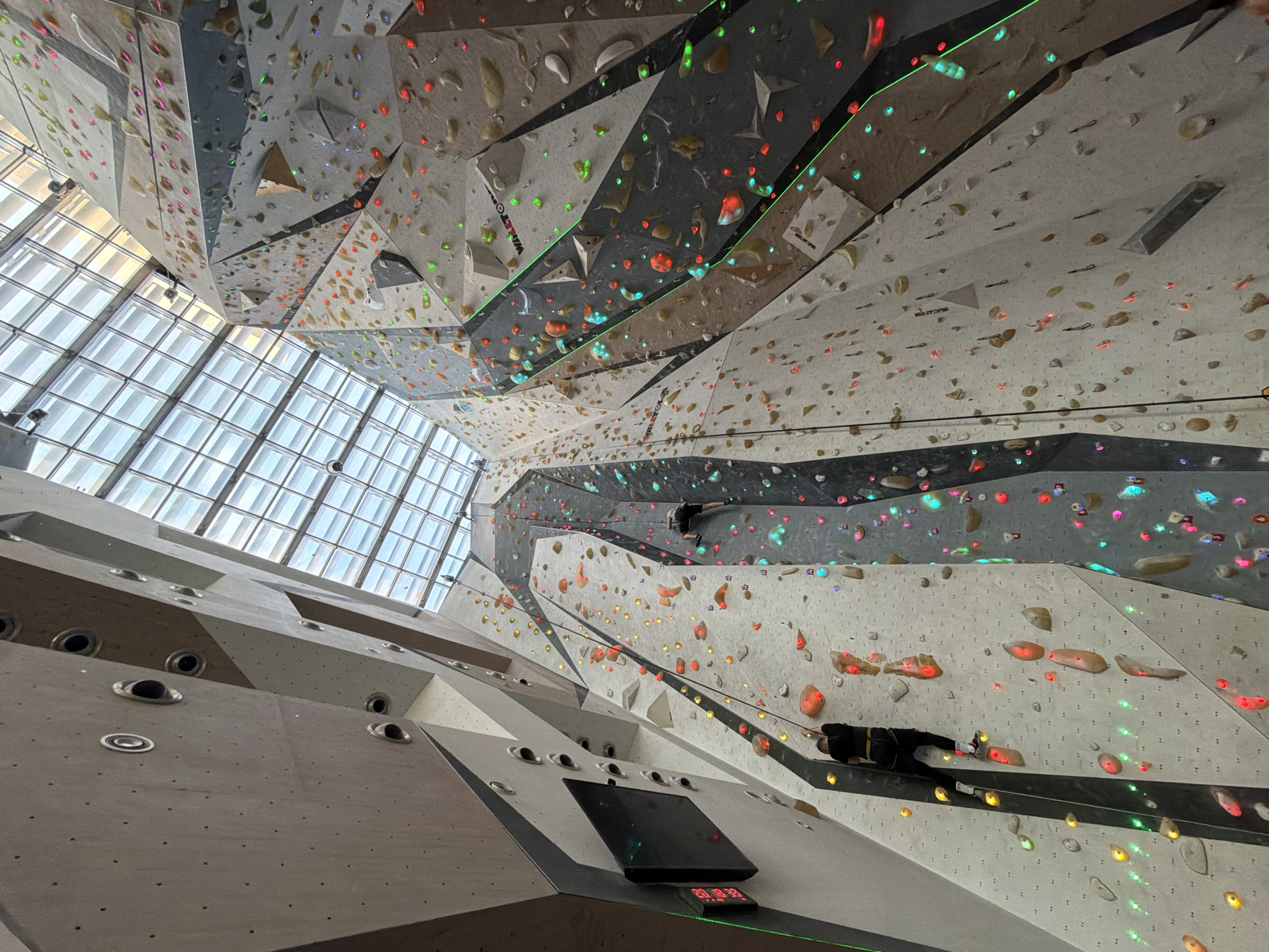 This is an interior shot of a modern, multi-story indoor climbing gym. The scene is dominated by a vast, geometrically paneled climbing wall system, featuring various angles and overhangs in shades of light grey, brown, and dark grey. A large, gridded window structure on the left side of the image allows natural light into the facility, suggesting it is daytime.
The climbing walls are densely covered with numerous climbing holds of various shapes and sizes. A distinctive feature is the integrated lighting system, with many holds glowing in bright colors like red, green, blue, and yellow, likely indicating specific climbing routes or interactive challenges.
At least two climbers are visible. One climber, dressed in dark clothing, is higher up on the right, partially obscured. Another climber, also in dark attire with some yellow accents, is actively engaged in climbing on a lower, brightly lit section of the wall to the right. Black auto-belay ropes run down sections of the wall, indicating safety equipment for climbers.
A digital display is integrated into a lower wall section, showing the time "20:18:32". The brand name "WALLTOPIA" is visible on several grey panels of the climbing structure, indicating the manufacturer of the climbing system. The overall atmosphere suggests an advanced and engaging climbing environment.