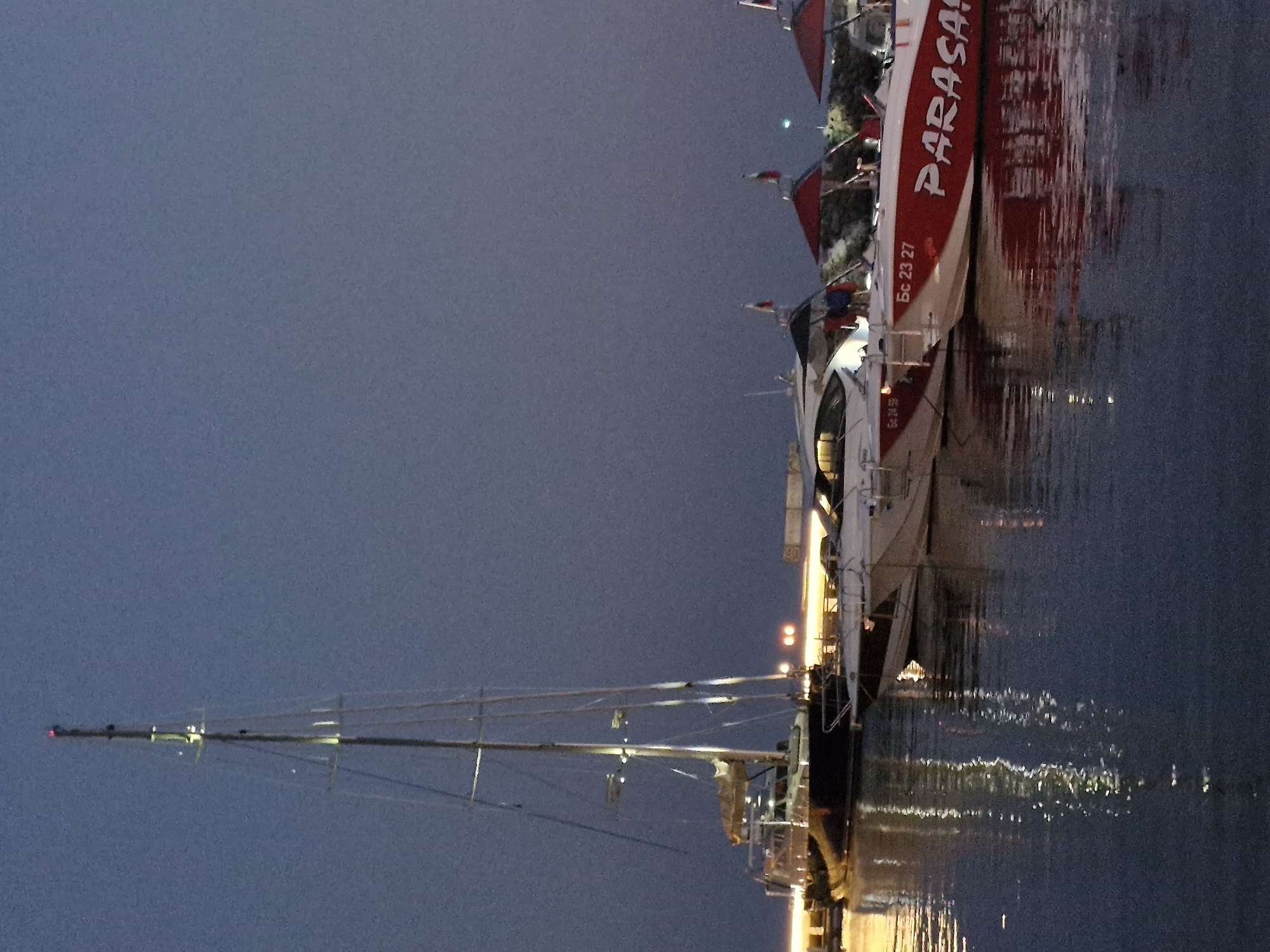 The image depicts a marina in Sveti Vlas, Bulgaria, at dusk. A tall sailboat with illuminated rigging is docked on the left. To its right, several smaller motorboats are moored. One of these motorboats has "PARASA" written in red lettering on its side. Red and black umbrellas are visible on some of the boats. The sky is a deep blue, and the water reflects the lights of the boats and the marina. There are no people clearly visible in the image. The overall scene suggests a peaceful evening at a coastal town.
