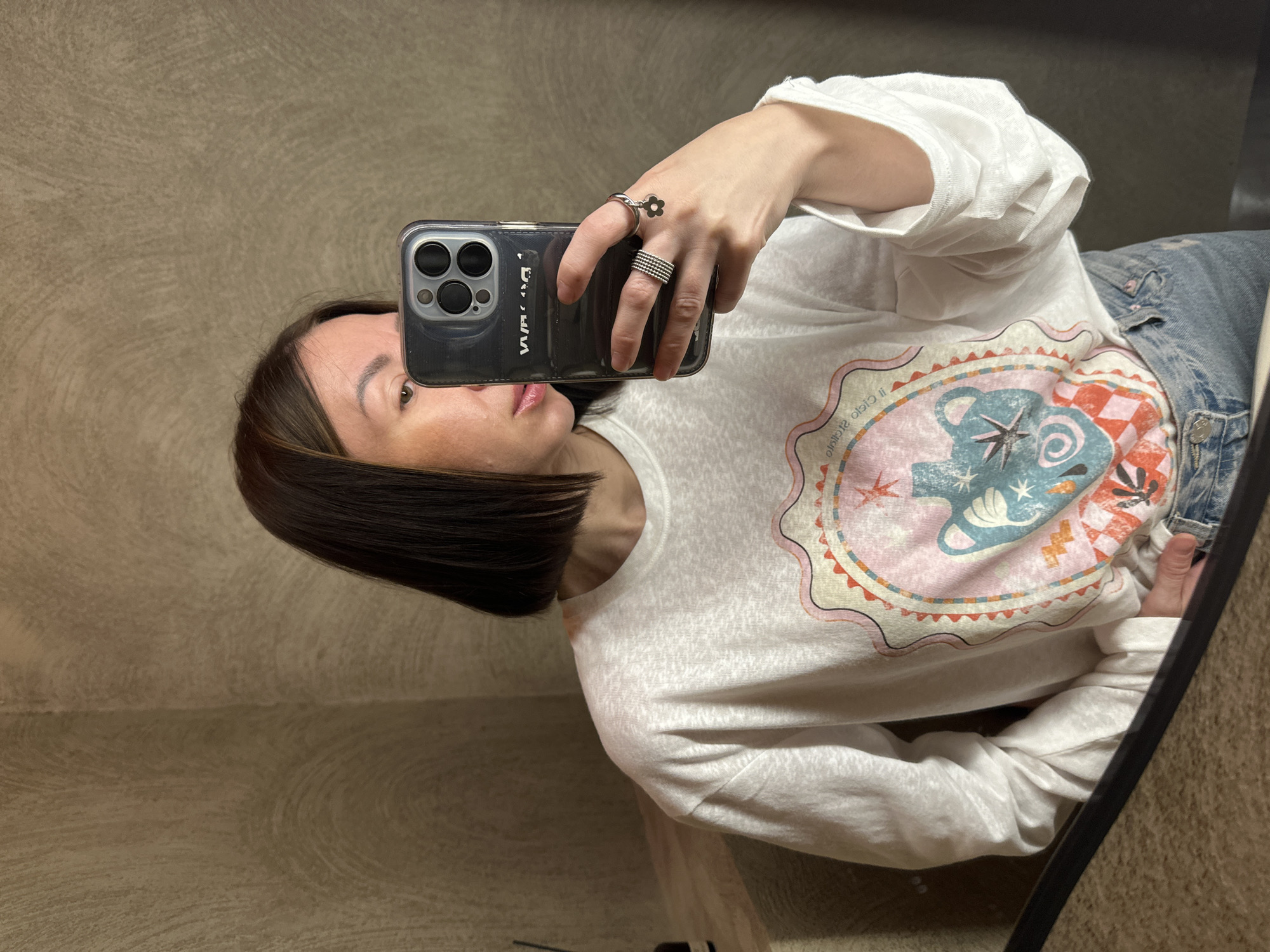 An adult female is captured in a mirror selfie within an indoor setting in Усады, Russia. She has dark, shoulder-length bob-style hair with subtle lighter highlights, and fair-to-medium skin tone with visible minor facial irregularities on the left cheek/forehead. Her lips are slightly parted, showing a natural or lightly glossed finish. She wears a loose-fitting, long-sleeved white t-shirt featuring a prominent circular graphic design. The graphic includes an urn-like vessel in shades of blue and teal, a starburst motif, and patterns in pink, orange, and red tones; reversed text, possibly "Cielo Olojo" or "Cielo d'Olio," is visible above the design. Light-wash denim jeans are partially visible below the shirt.
The subject holds a dark-cased smartphone (appears to be an iPhone with a triple-lens camera array) in her left hand, positioning it to capture her reflection. The phone case has reversed text, possibly "VVAA" or "DVVA." On her left hand, she wears two rings: a wide, multi-banded silver ring on the ring finger, and a single silver band with a small, dangling flower charm on the middle finger.
The background consists of a textured wall, finished in a light brown or greige color with subtle, swirling plaster patterns. A corner of the room is visible. To the upper left, a small section of a light-colored wooden shelf or counter is present, holding a dark, slender bottle, possibly a diffuser. The bottom of the image shows a dark, horizontal line, indicative of the edge of the mirror frame or a counter surface reflecting the subject's lower body. The lighting is diffused and even, with no harsh shadows.