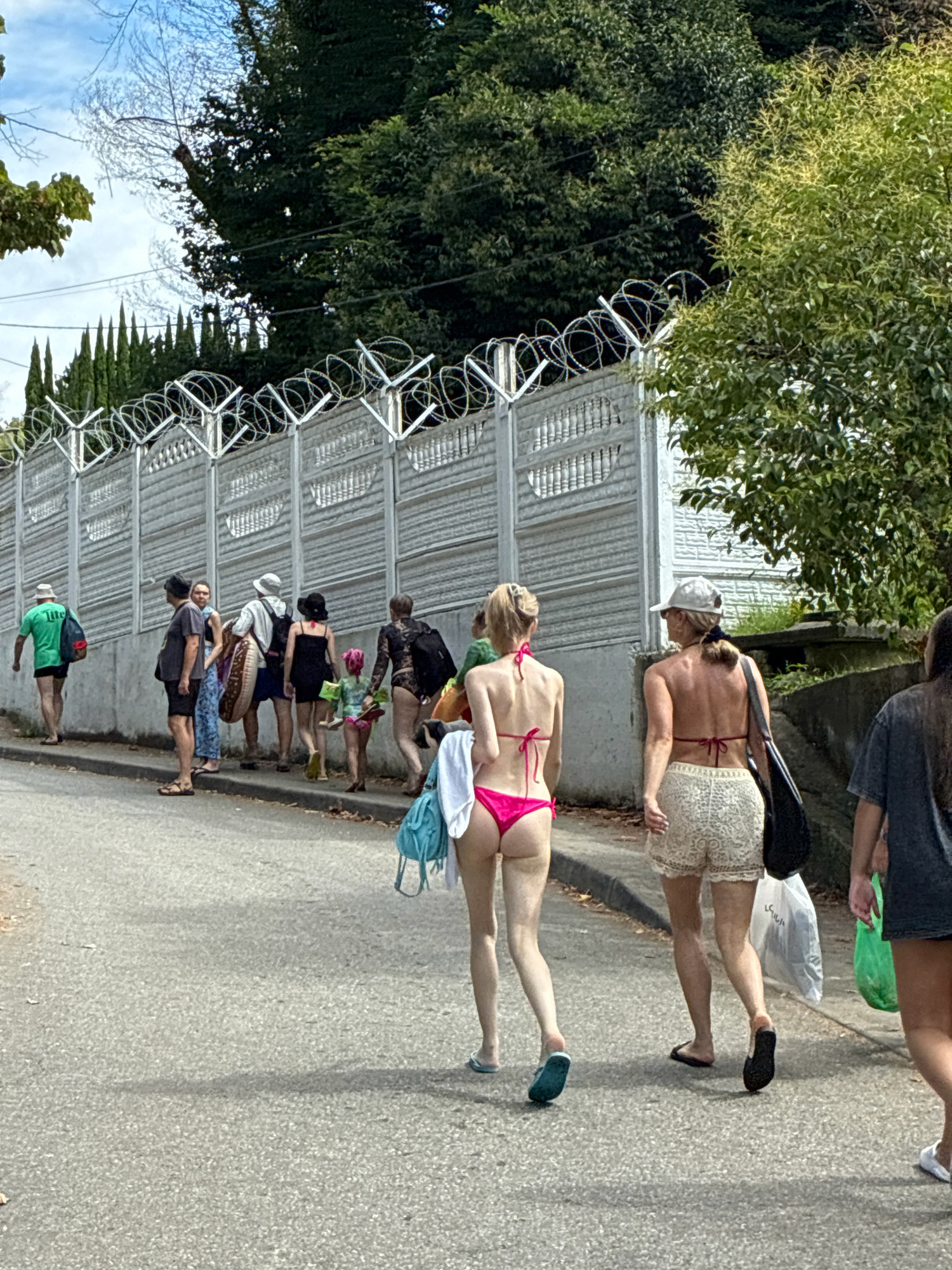 A group of people, some wearing swimwear, walk along a paved street in Sochi, Russia. The street slopes upwards and is bordered by a concrete wall topped with barbed wire. Behind the wall, lush green trees and foliage are visible, suggesting a warm climate. The sky is partially visible, showing blue patches and clouds. The people are diverse in age and attire, with some carrying bags and beach accessories, indicating they are likely heading towards or returning from a beach or recreational area.