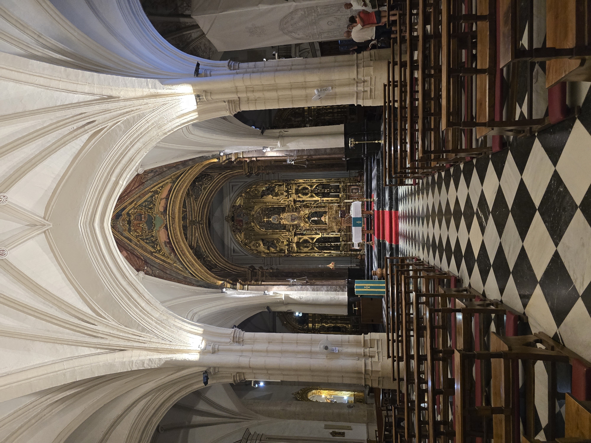 This is a photograph of the interior of the Jaén Cathedral, located in Jaén, Spain. The image shows the nave of the cathedral, looking towards the altar. The architecture features high vaulted ceilings with ribbed arches supported by stone columns. The floor is a black and white checkered marble pattern. Rows of wooden pews with red cushions line both sides of the nave. In the distance, the ornate altar is visible, adorned with golden decorations and religious iconography. A red carpet leads up to the altar. Several people are standing near the altar area, observing the scene. The lighting in the cathedral appears to be artificial, with spotlights illuminating the architectural features and the altar.