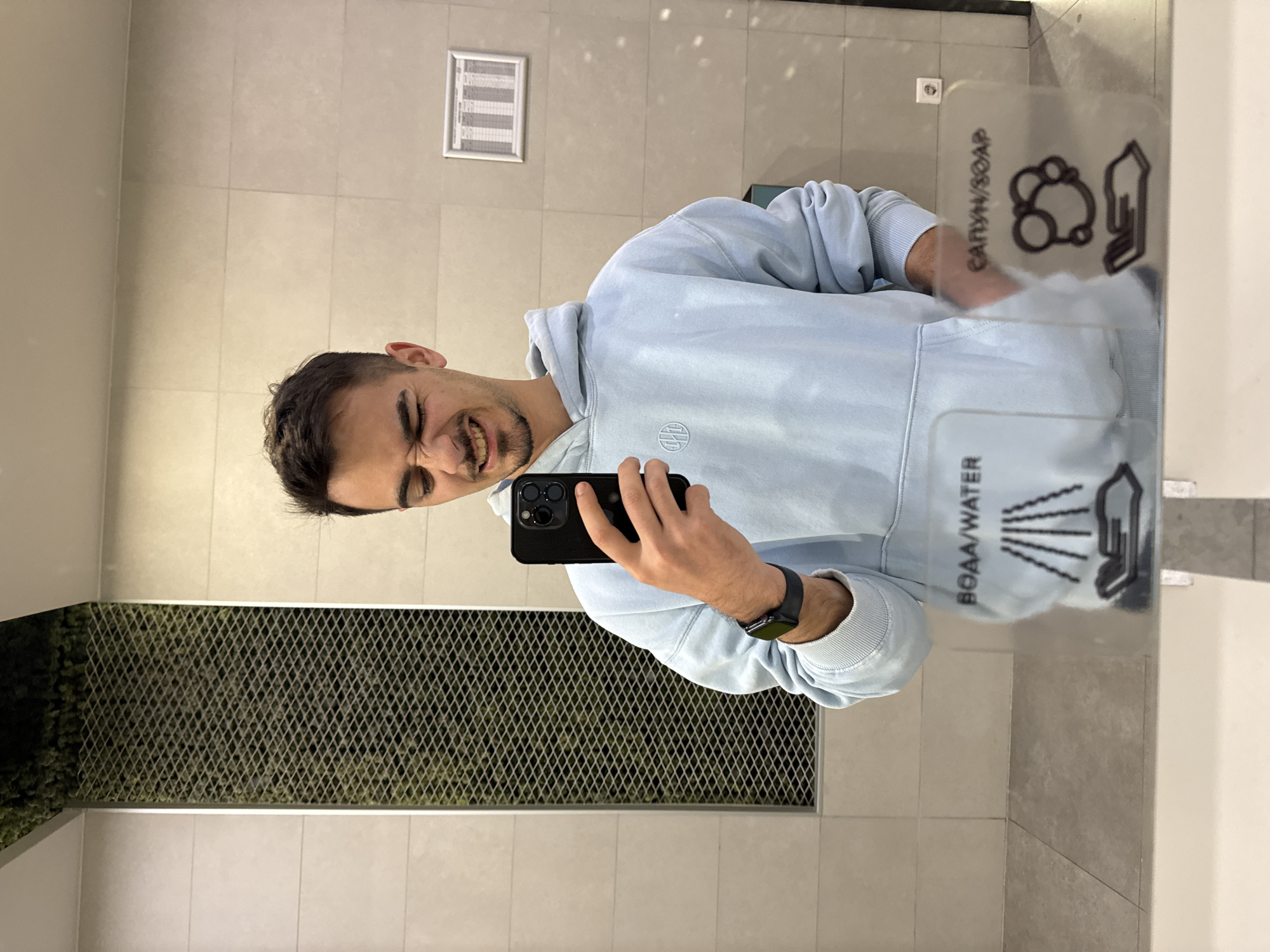 The image, rotated 90 degrees clockwise, captures a young man taking a mirror selfie in what appears to be a modern public washroom or similar indoor facility in Sofia, Bulgaria. The man, with short dark hair and a beard, is making a humorous, exaggerated facial expression, squinting his eyes and revealing a wide grin. He is dressed in a light blue hoodie, which has a small, stylized circular logo on its chest, and wears a black smartwatch on his left wrist. He holds a black smartphone with multiple camera lenses to capture the image.
In the foreground, a transparent sign or dispenser is prominent. It displays icons for hygiene, specifically "САПУН/SOAP" (bubbles and a hand) and "ВОДА/WATER" (water splashes and a hand), with the text presented in both Cyrillic and English. The background features light-colored tiled walls, suggesting a clean and functional interior. To the left, a decorative panel composed of a metal mesh layered over greenery, possibly artificial, adds a design element. A rectangular ventilation grille and an electrical outlet are also visible on the wall. The overall mood conveyed is lighthearted and playful.