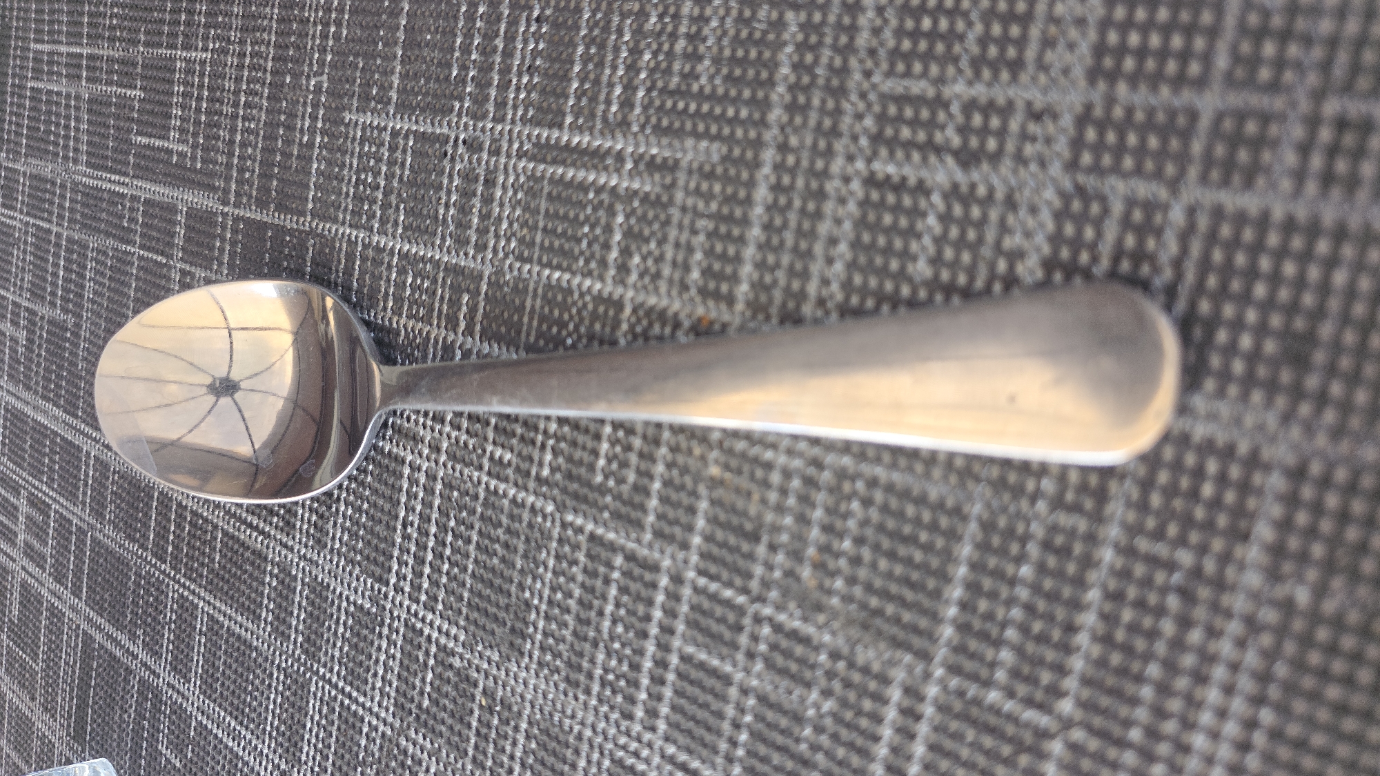 A metal spoon is placed on a textured, grey and white checkered surface. The spoon is positioned vertically in the center of the frame, with its bowl facing upwards and reflecting a pattern of lines and circles. The handle of the spoon extends downwards and out of view. The surface beneath the spoon appears to be a placemat or tablecloth. No people or any other objects are visible. The image does not contain any information or context that would allow for the determination of its location in the city of Hamm, Germany.