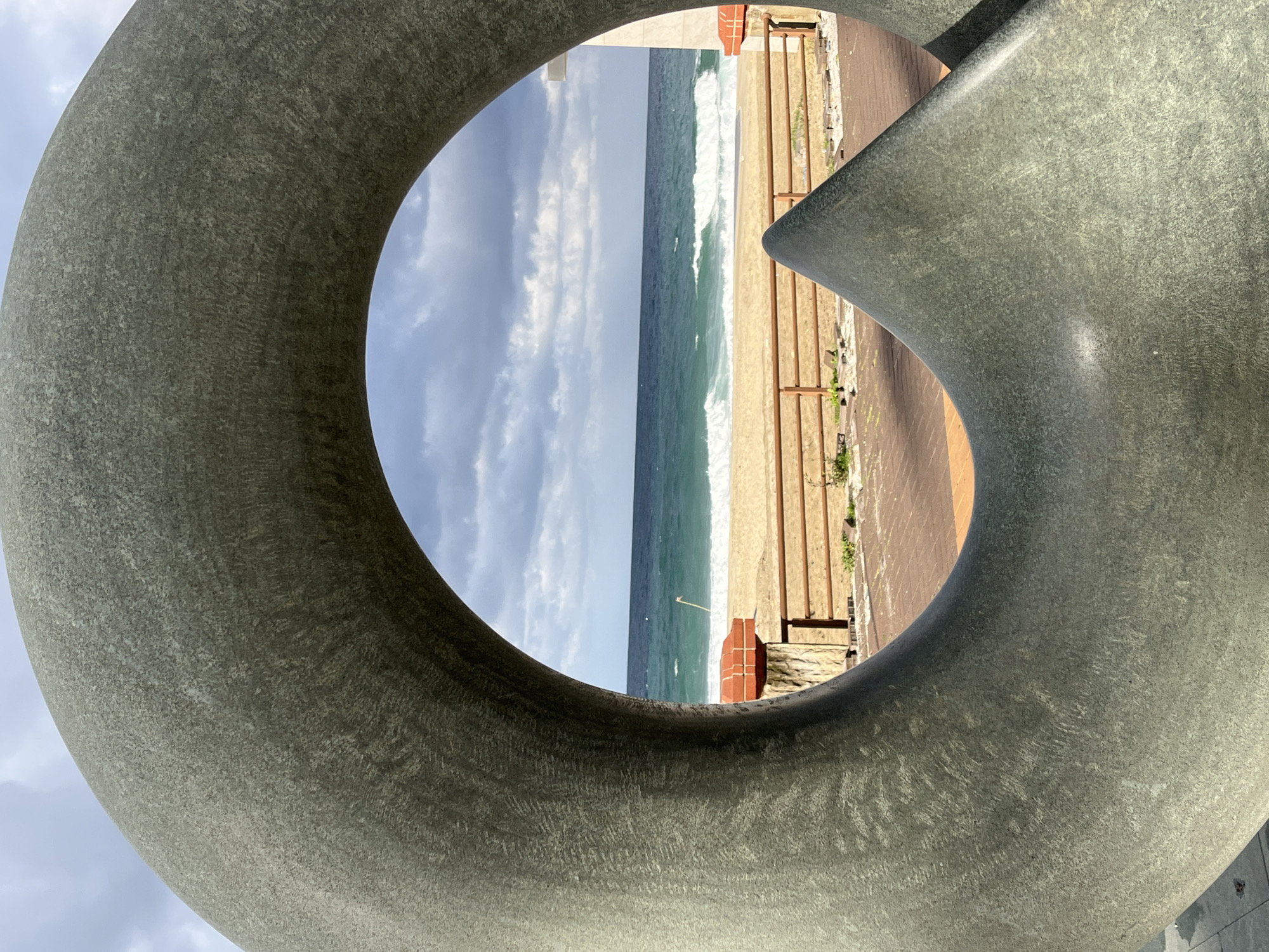 The image depicts a view of a beach through a large, circular sculpture made of a gray, textured material. The beach is sandy with waves breaking. A wooden boardwalk runs parallel to the beach, separated by a rusty metal railing. Beyond the beach, the sky is partially cloudy. The location is Alanya, Turkey.