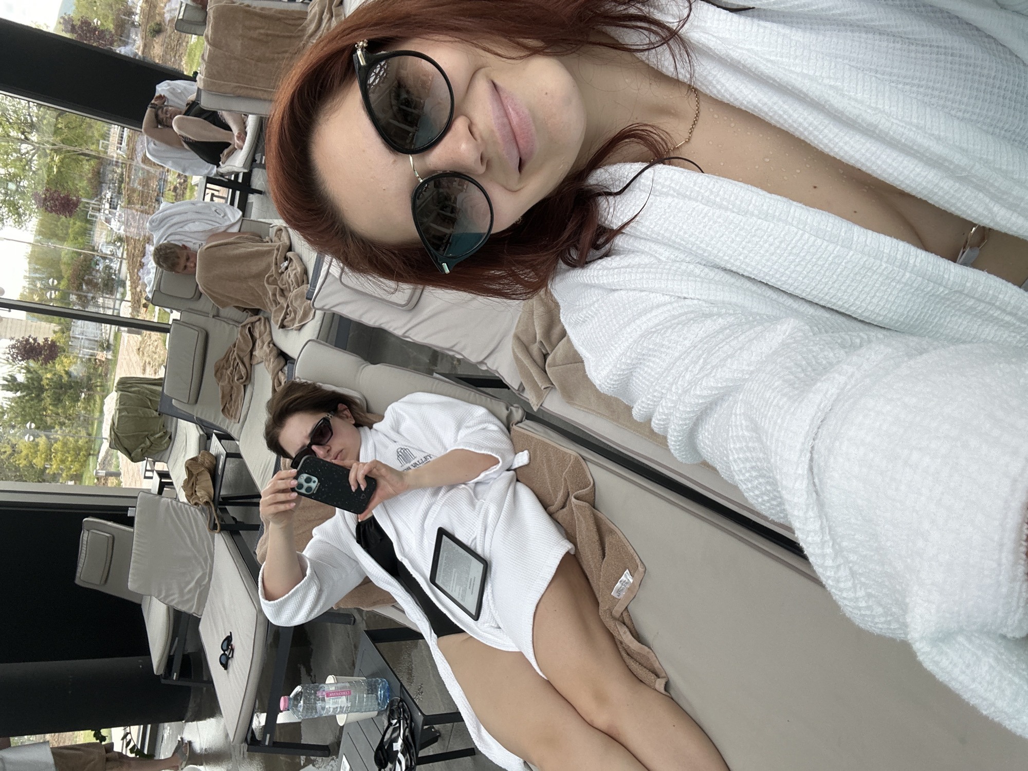A woman wearing sunglasses and a white robe is lying on a lounge chair in what appears to be a spa or resort. She is taking a selfie with her head tilted. The white robe is open, revealing her bare chest and wet skin. Other individuals are reclining on lounge chairs in the background. Another woman wearing sunglasses and a white robe is lying on a lounge chair in the foreground and holding a smartphone. An e-reader sits on top of her legs. A bottle of water sits on a table near her lounge chair. The location appears to be the "Shipton Valley" resort, as imprinted on the white robe of the woman holding the smartphone. The exterior features a view of greenery and buildings. The location is in Kazanlak, Bulgaria.