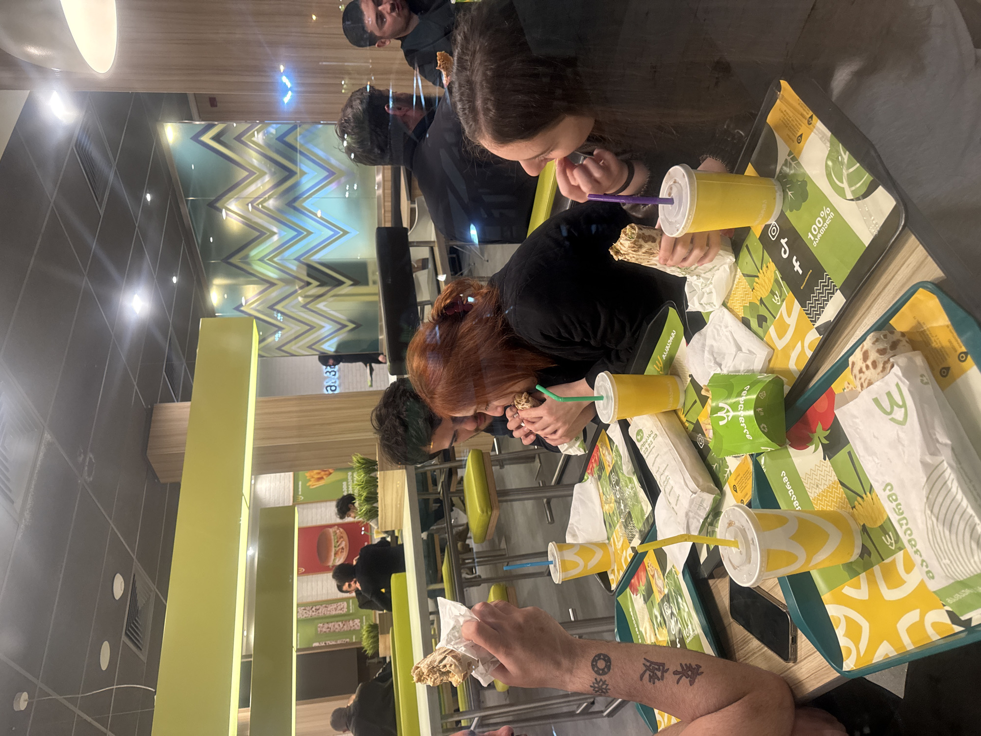**Scene:** An indoor fast-food restaurant, possibly located in Tbilisi, Georgia.
**People:** Several individuals are seated at a table, eating and drinking. At least two individuals have visible tattoos.
**Objects:** Drinks with straws, wrapped food items (possibly shawarma), trays with designs featuring the Georgian language.
**Actions:** People eating, drinking, and interacting.
**Interactions:** Individuals are gathered around the table, focused on consuming their meals.
**Environment:** The restaurant interior includes a colorful geometric design on the wall, and the layout suggests a casual dining setting.