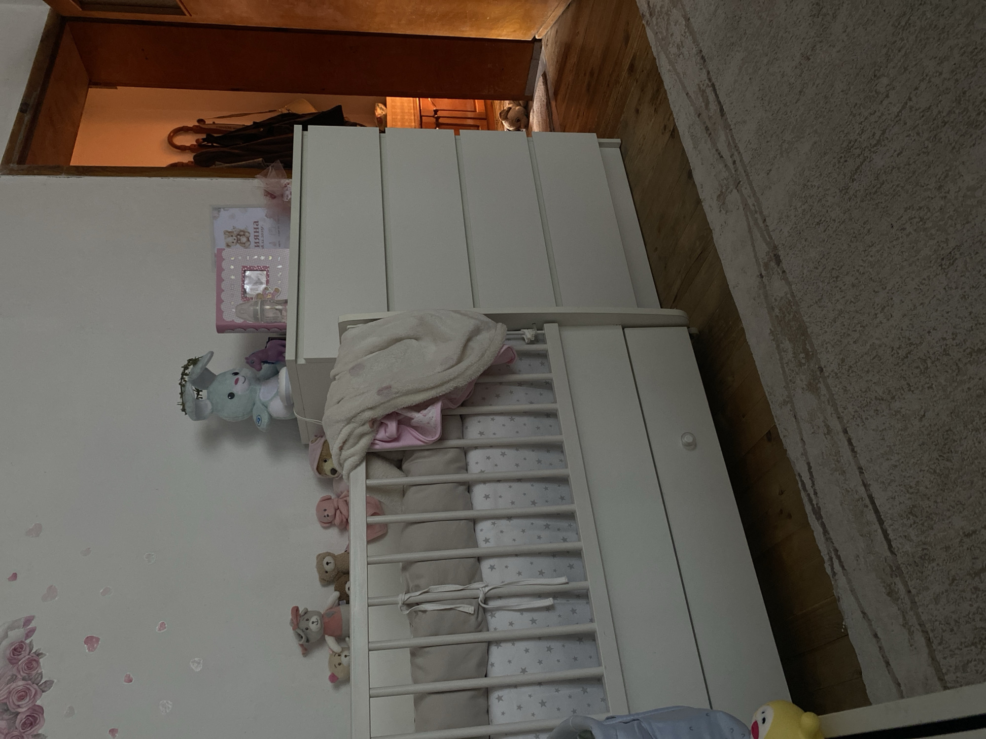 The image displays an indoor scene, likely a child's nursery or bedroom. The room features white walls, wooden flooring, and a light-colored, textured area rug.
A prominent white wooden crib stands in the center-right of the frame, equipped with vertical slats. Inside, there are soft grey bumper pads secured with white ties, and white bedding adorned with small grey stars. A light pink blanket is draped over one side of the crib. Several small plush toys, including teddy bears and rabbit-like figures, are attached to the crib rails. Another larger light blue plush toy is hung on the wall above the crib. A yellow toy is partially visible at the bottom right.
To the left of the crib, a white cabinet or dresser with multiple sections stands. On top of it are two decorated books or photo albums; one pink with a small mirror, and another white featuring a teddy bear design. Visible text on one album reads "ИЯНА" and "К.Б. 12. 2022Г.", suggesting a name and a date.
The white wall to the left is decorated with a delicate decal of pink roses and scattered pink petals. In the background, an open doorway or archway reveals a warmly lit adjacent area with wooden paneling. A folded dark umbrella with a curved wooden handle is placed within this opening. Notably, a small, light-colored animal, possibly a dog or cat, is peeking into the room from this background area.
The room appears tidy and well-appointed for a baby or small child, creating a serene and gentle atmosphere. No human activity is depicted. The lighting suggests an indoor setting, possibly with soft ambient light. While the location context of Blagoevgrad, Bulgaria, is provided, no specific visual cues in the image confirm this.
