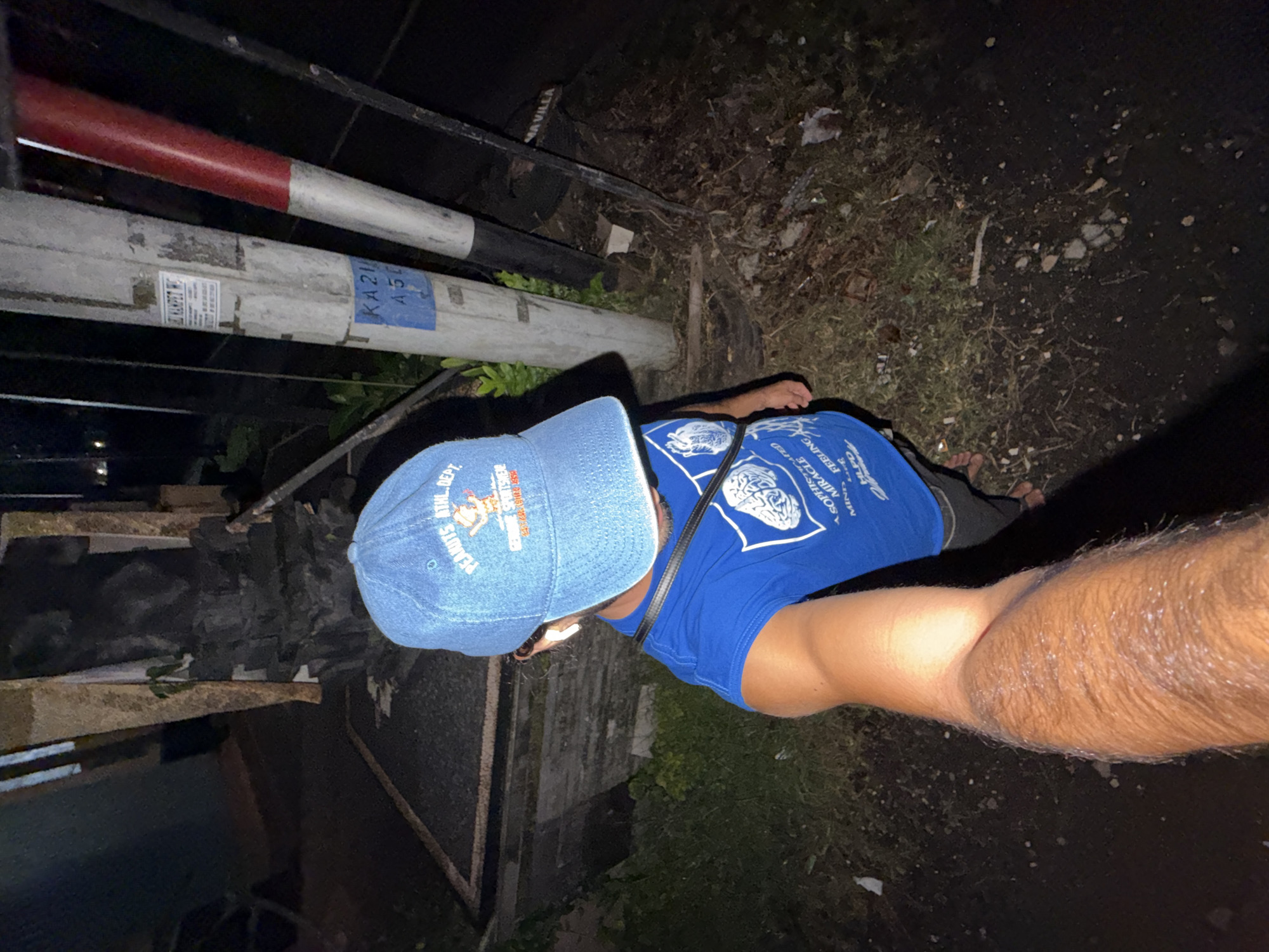 A selfie taken at night in North Kuta, Indonesia. The subject, wearing a blue "Peanuts" baseball cap and a blue t-shirt with a brain logo, is standing next to a utility pole marked "KA21". The surroundings include a dark, possibly paved area with some vegetation and an adjacent stone wall.