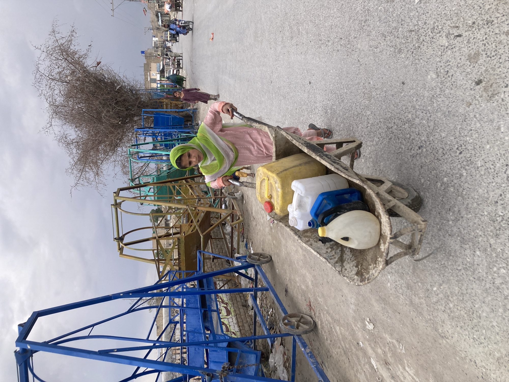 The image shows a young woman pulling a wheelbarrow filled with plastic containers, possibly for water, in Quetta City Tehsil, Pakistan. She is wearing a pink outfit and a green headscarf. The surroundings show a dusty road, colorful metal structures that appear to be part of children's rides or amusement structures, and other people visible in the background.